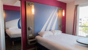 A hotel room with a bed, a bedside table, a mirror, and a window with a view of buildings.
