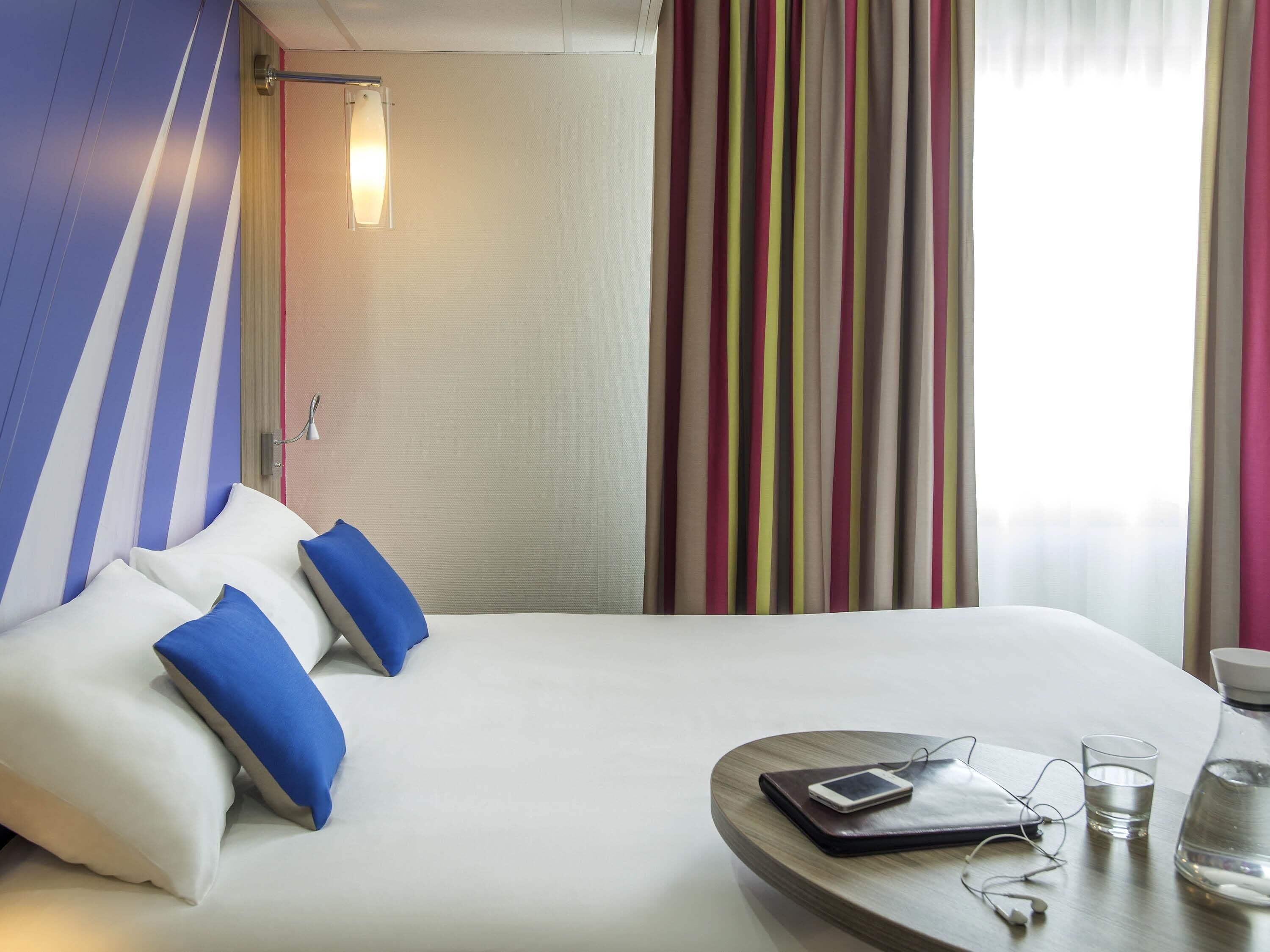 A hotel room with a bed, pillows, a bedside table with a phone, glasses, and a water bottle.