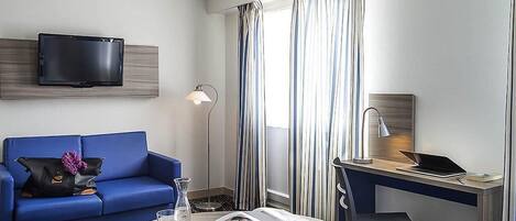 A hotel room with a blue sofa, a round coffee table with a book and glass, a desk with a laptop, a TV mounted on the wall, and striped curtains.