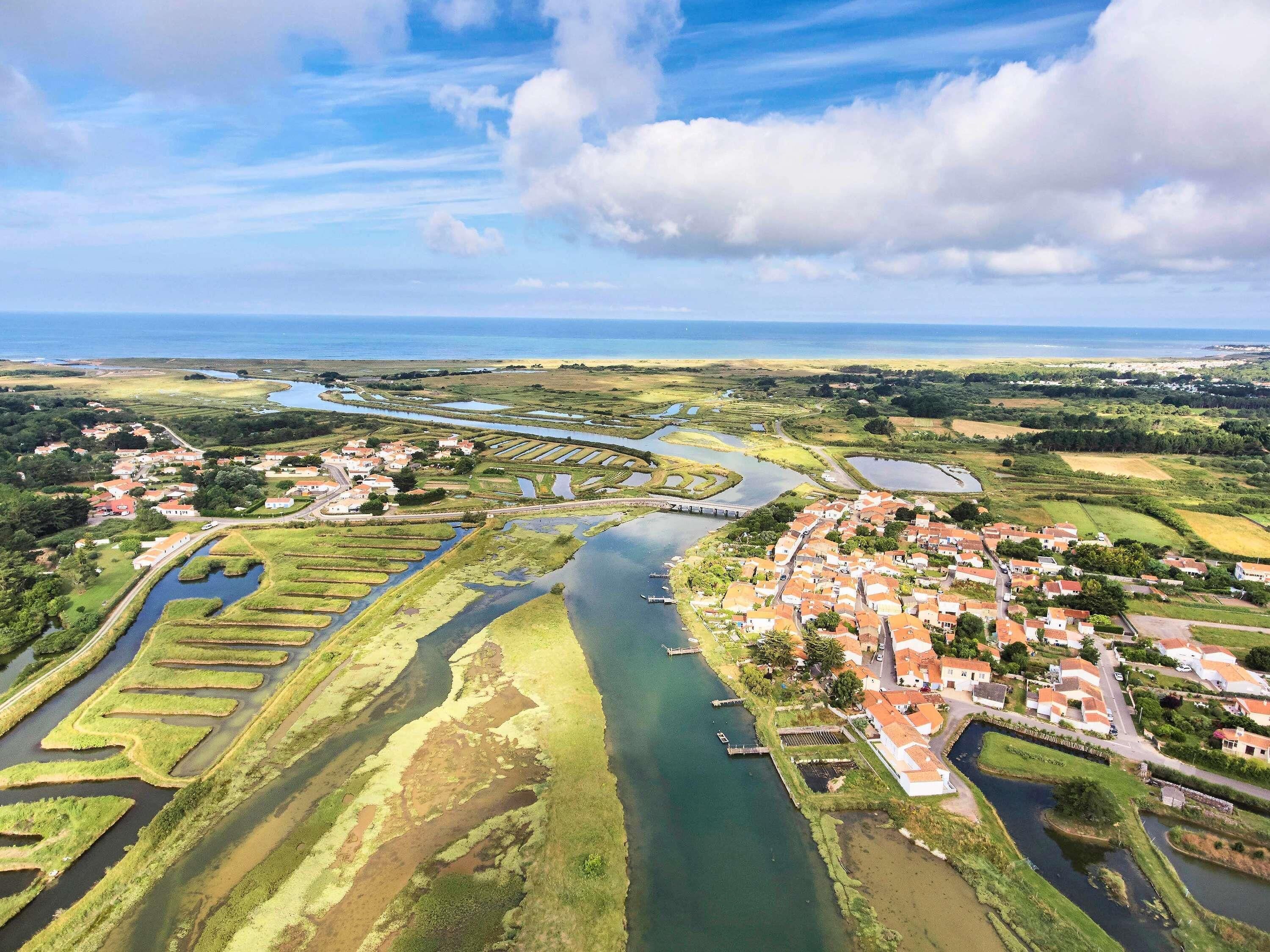 An aerial view of a coastal town with a river, green fields, and a bridge connecting to the mainland.