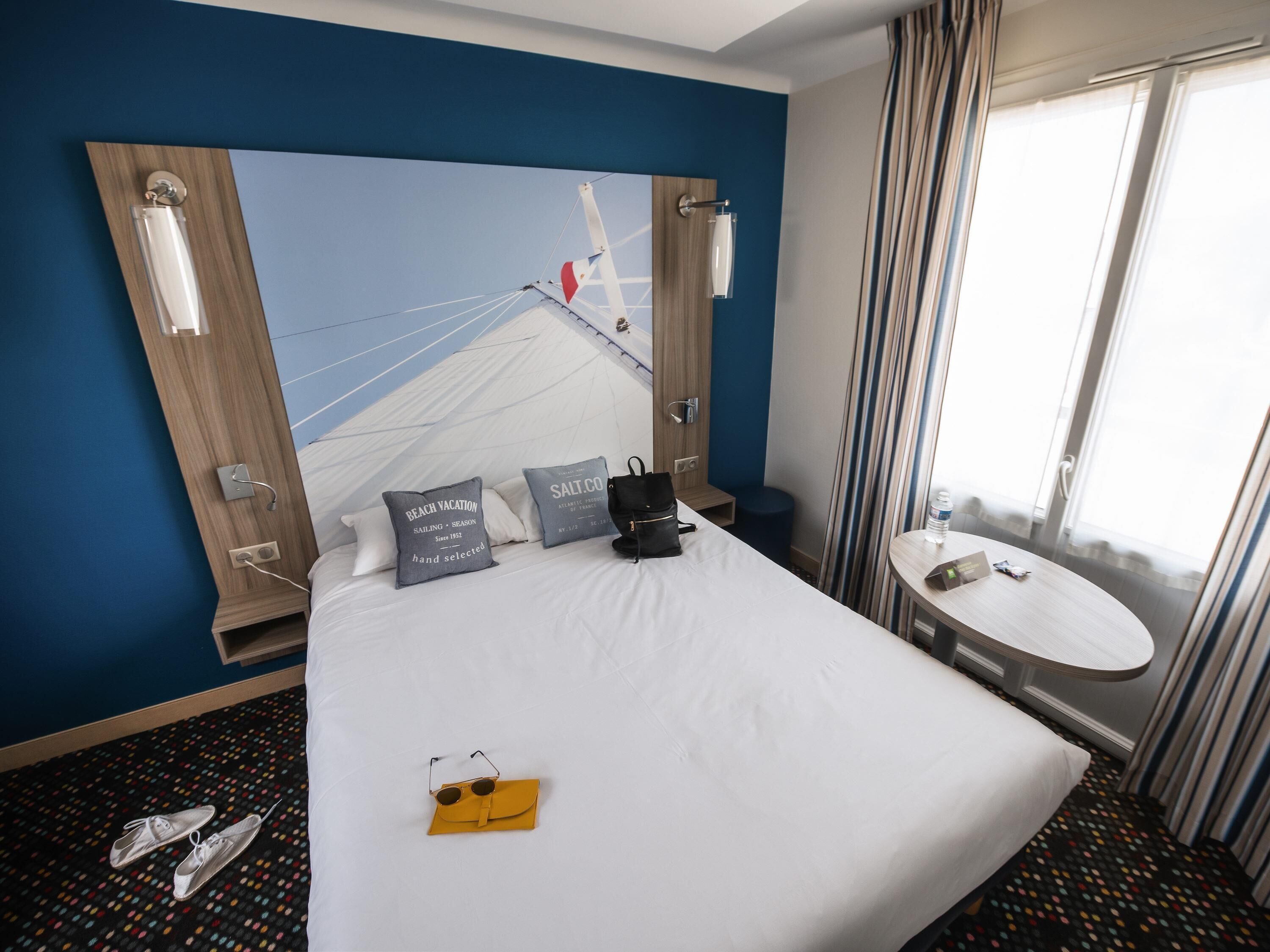 A hotel room with a large bed, a small round table, a window with curtains, and a wall-mounted mirror.