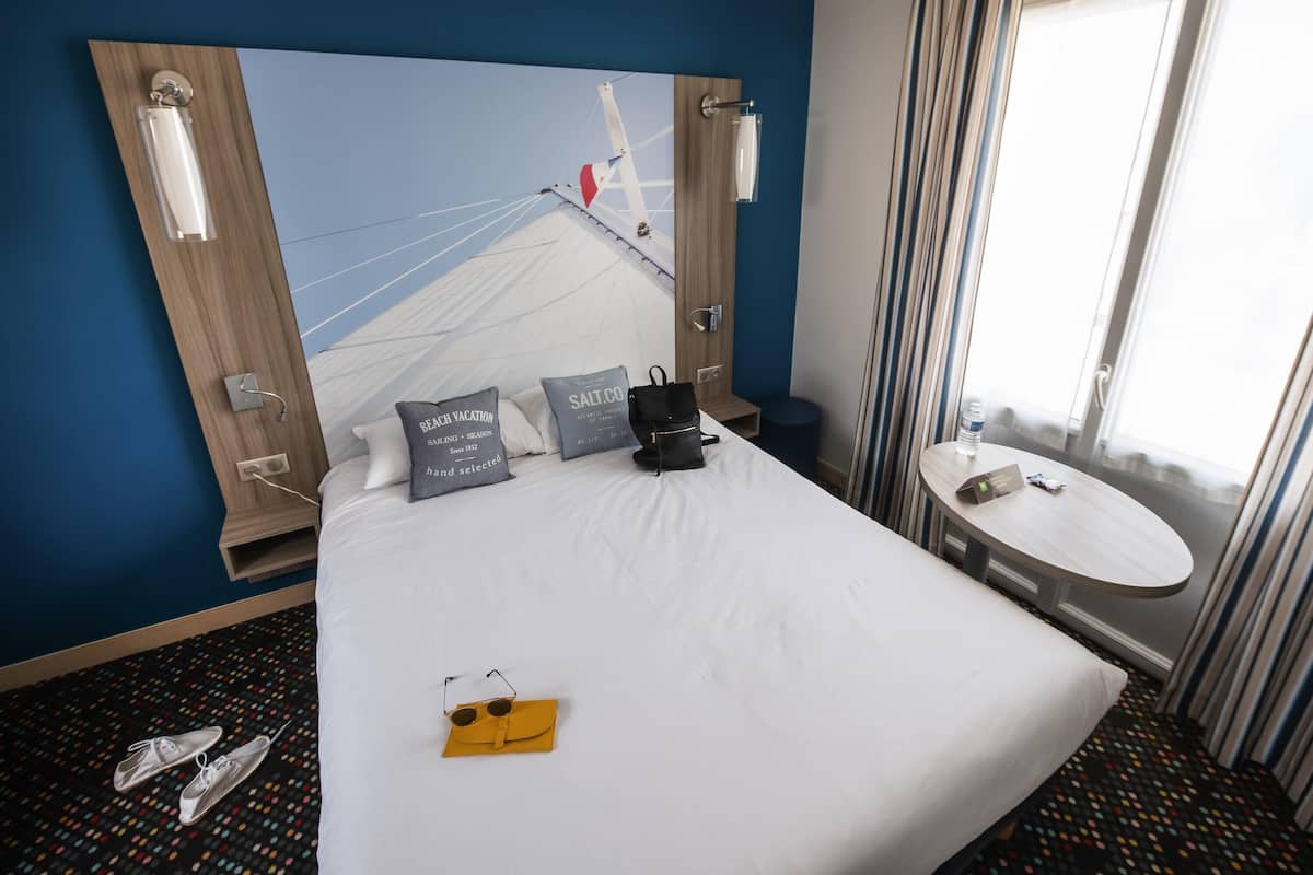 Une chambre d’hôtel avec un grand lit, une petite table ronde, une fenêtre avec des rideaux et un miroir fixé au mur.