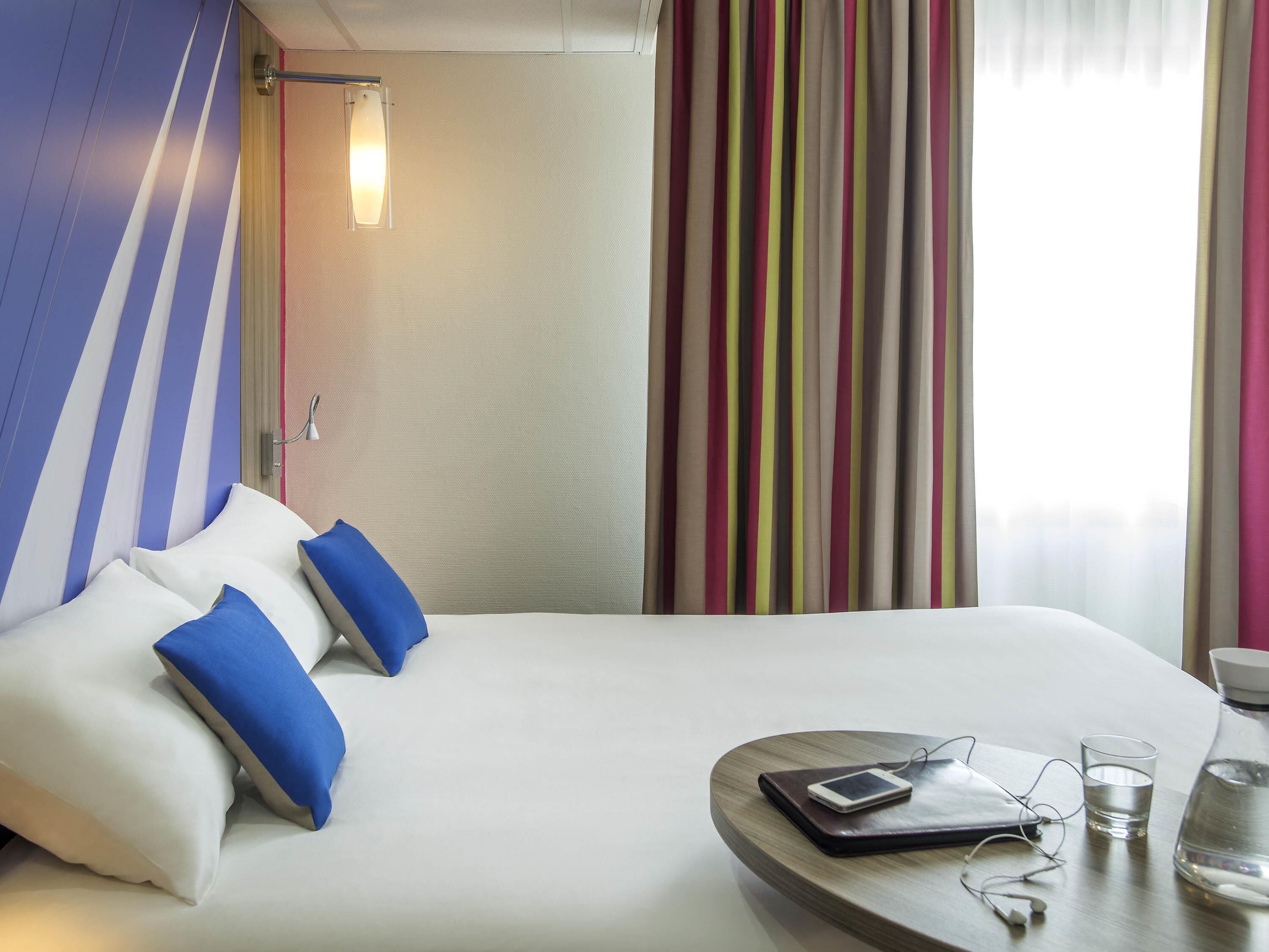 A hotel room with a bed, pillows, a bedside table with a phone, glasses, and a water bottle.