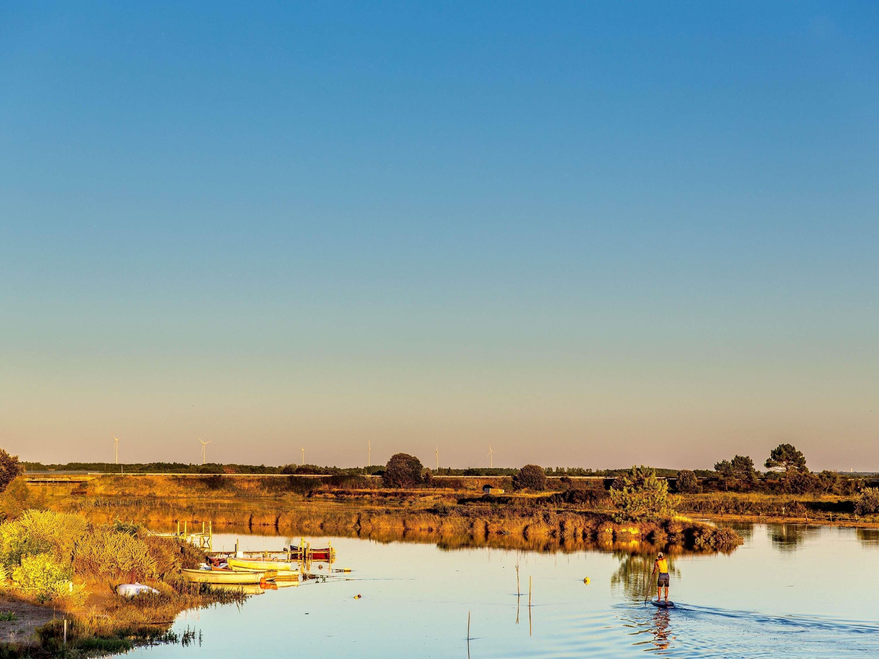 A calm water body with boats, a person standing by the water, and wind turbines in the background.