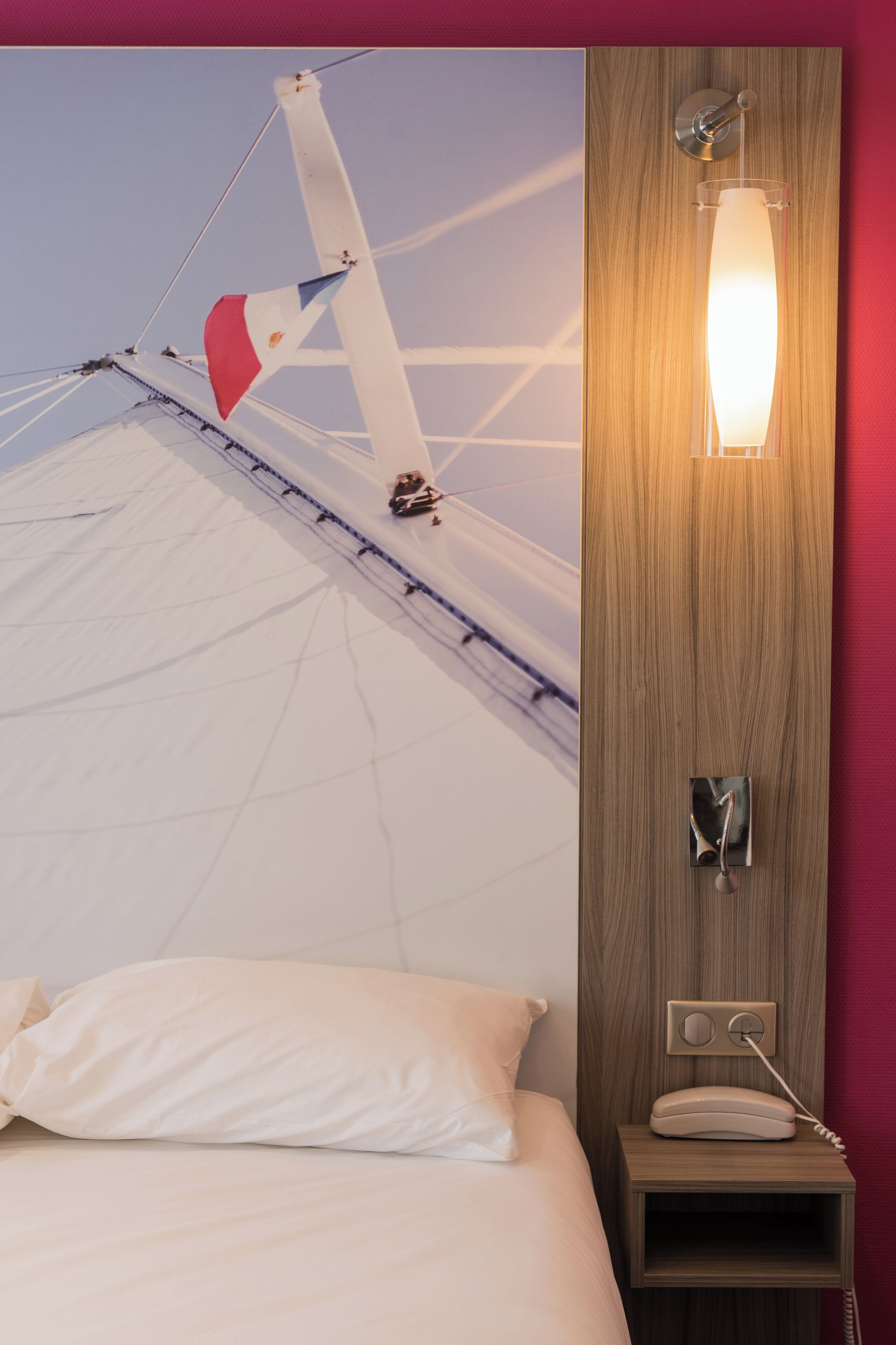 A hotel room with a bed, a wooden nightstand with a telephone, and a wall-mounted light fixture.
