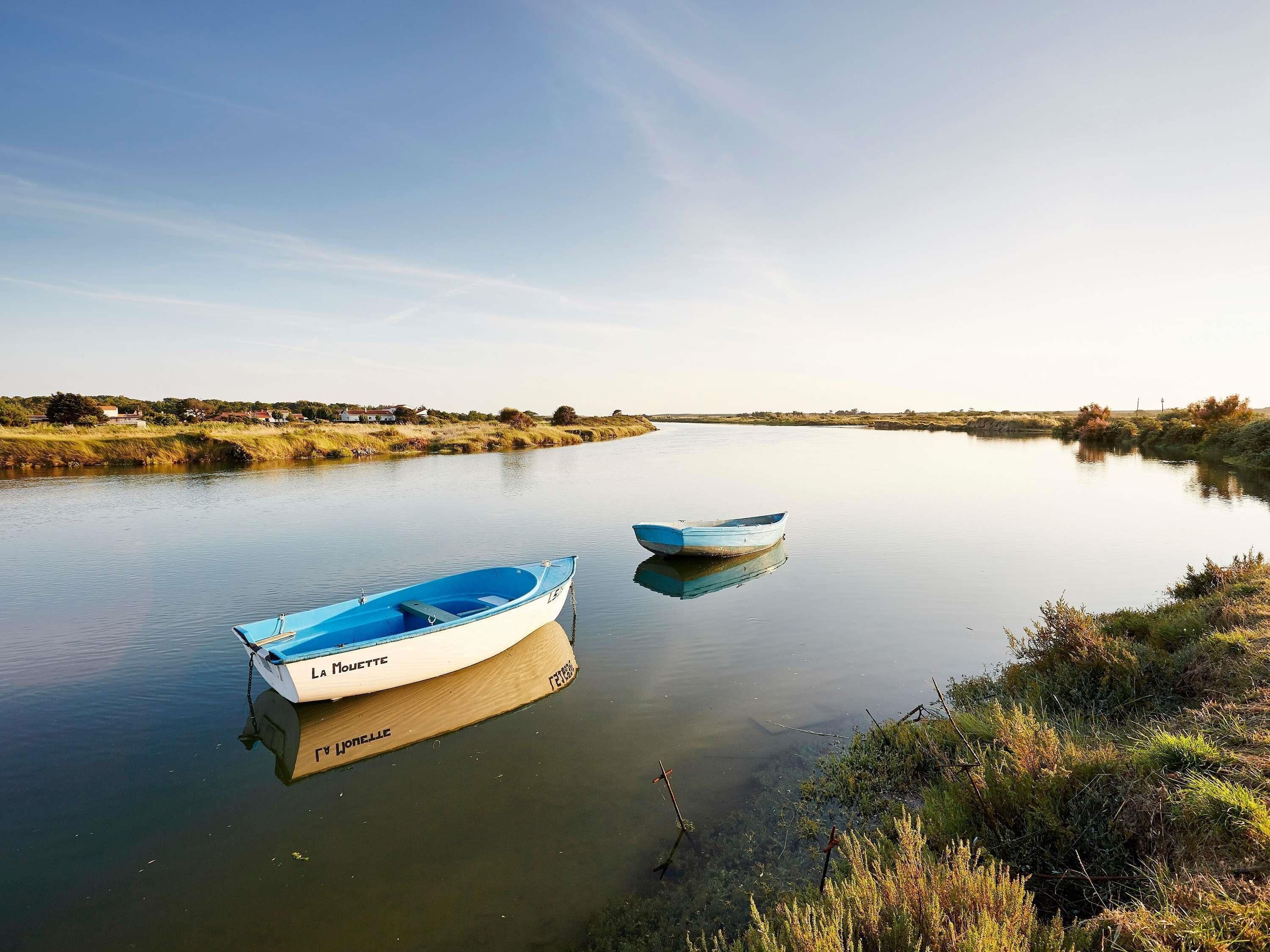 Deux bateaux sur une rivière paisible, bordée de verdure, avec des maisons visibles au loin.