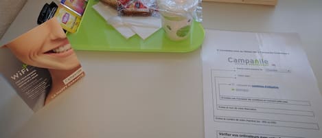 Zona de recepción de un hotel con una bandeja verde que contiene una taza blanca, una botella de agua y un paquete de snacks. Hay un cartel de WiFi y una tarjeta de bienvenida sobre la mesa.