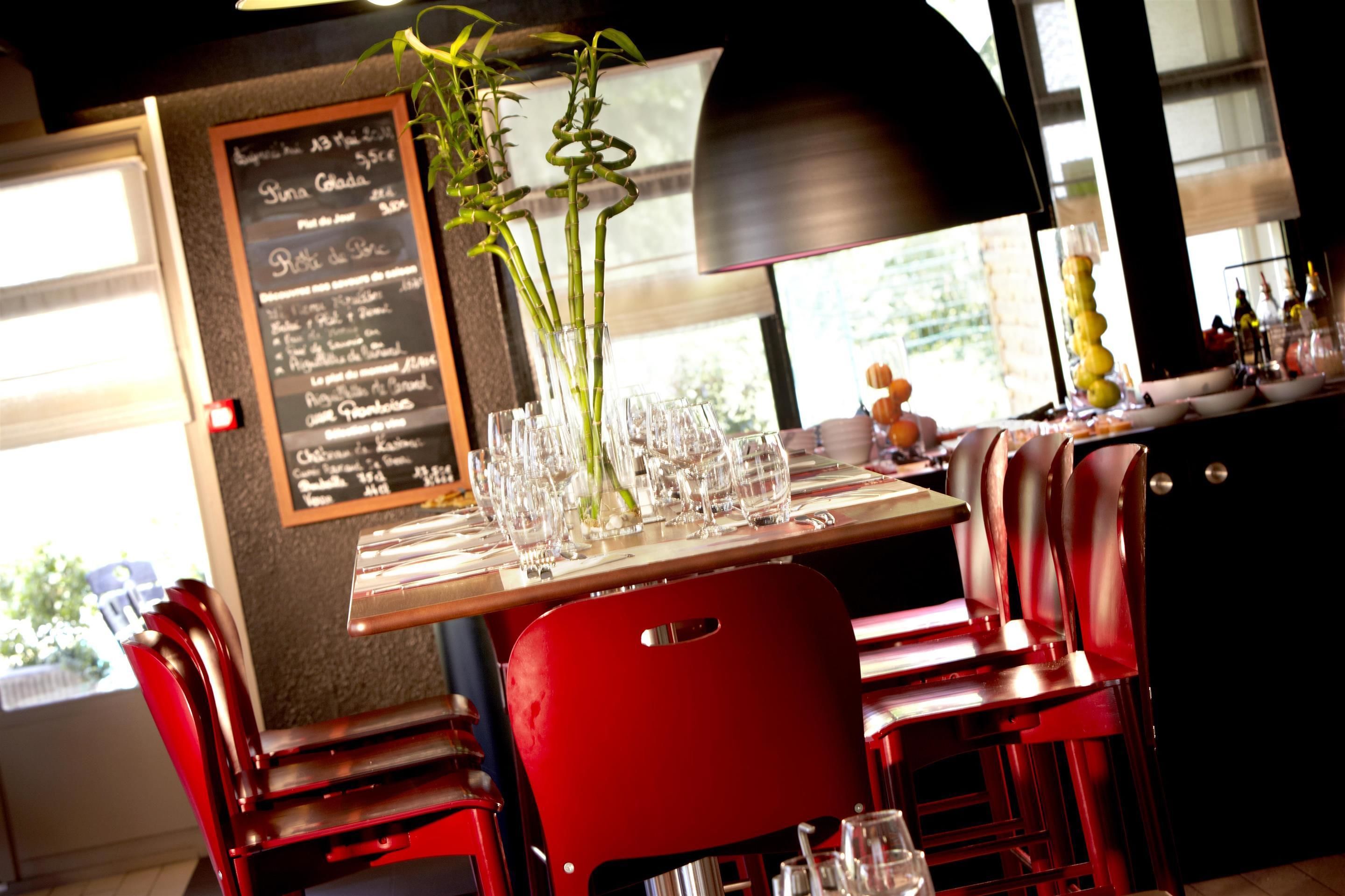 A restaurant interior with a wooden table set for dining, red chairs, a menu board, and a plant in a glass vase.