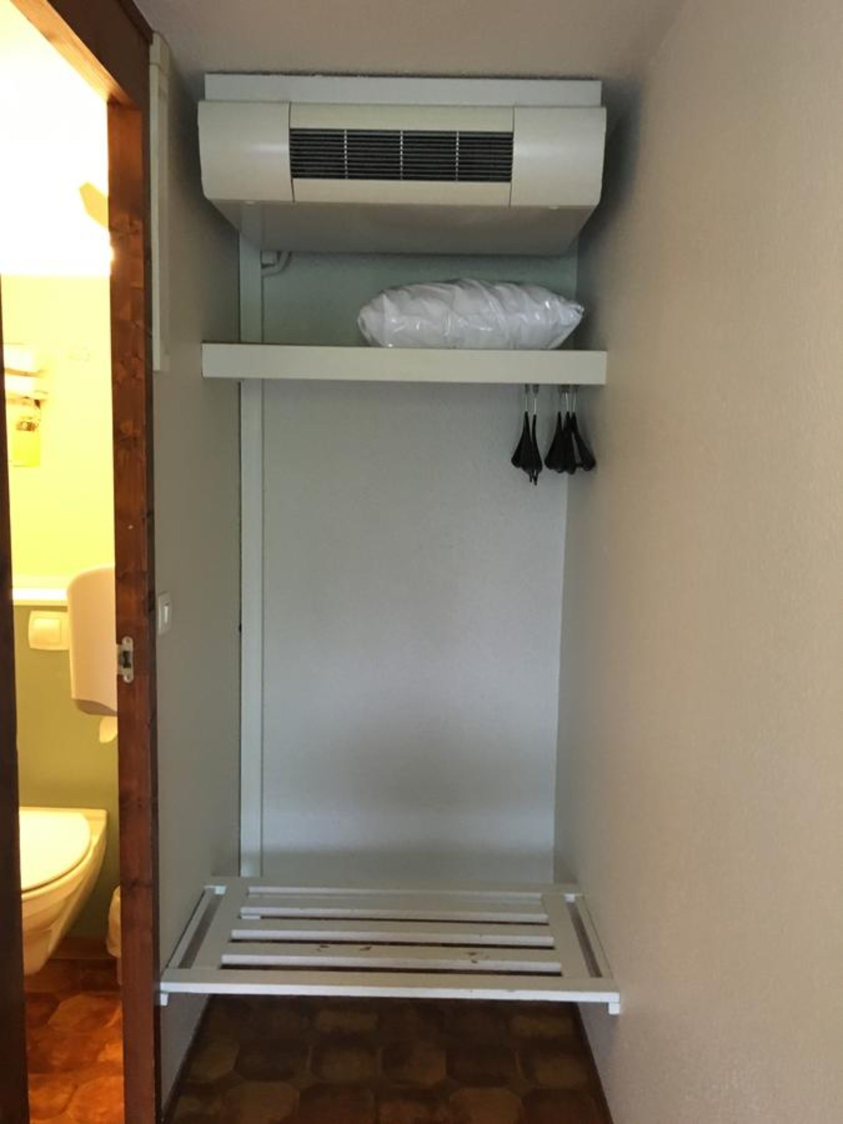 A compact, white-framed walk-in closet with a built-in air conditioning unit, a shelf with a plastic bag, and a hanging rack with black items.
