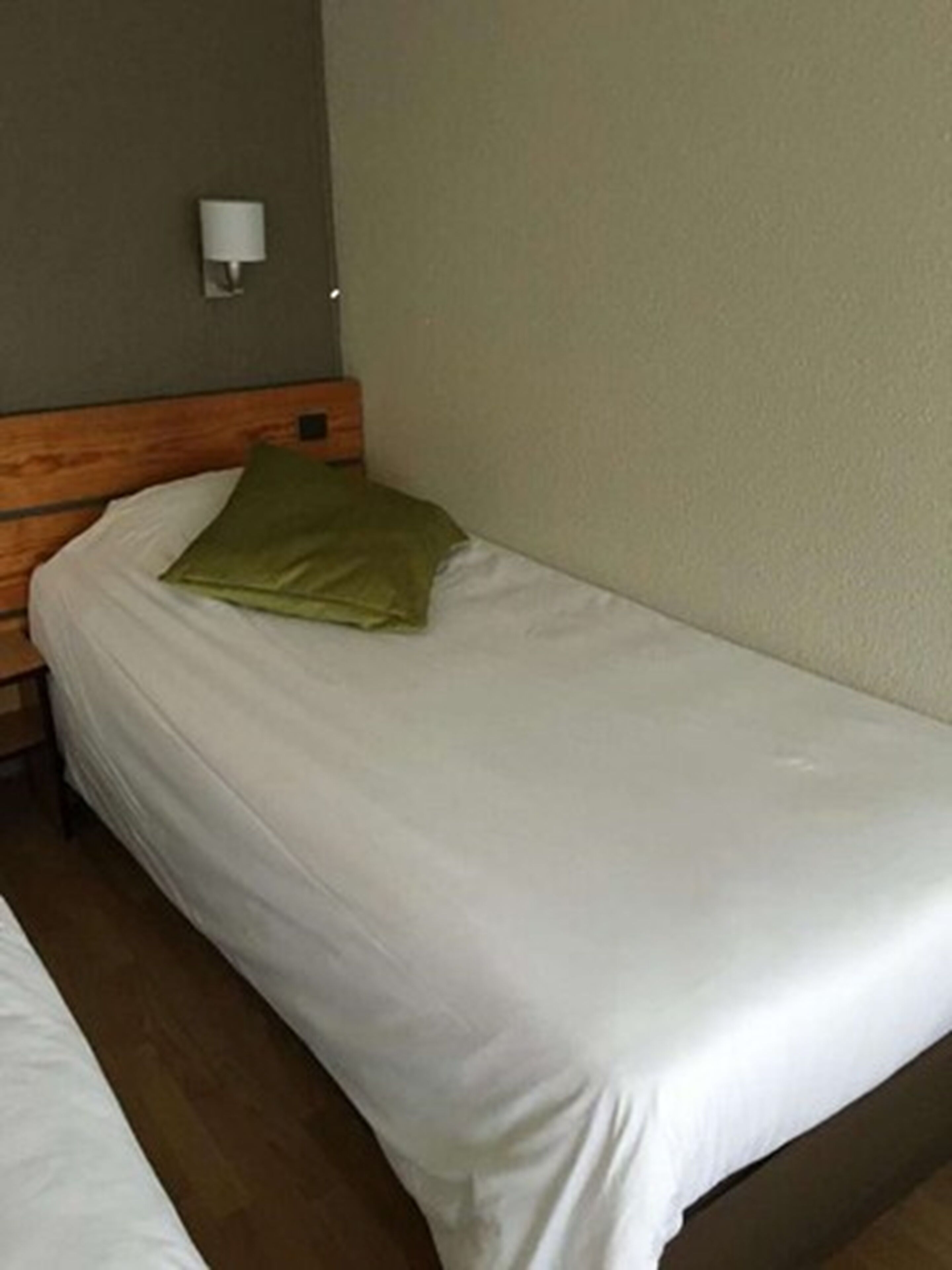 A single bed with a wooden headboard, a green pillow, and a white wall-mounted light fixture.