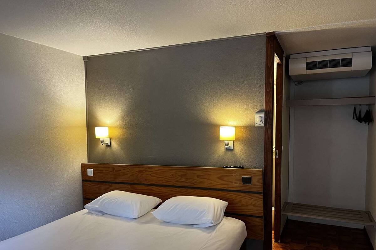 Habitación de hotel con cabecera de madera, dos lámparas de noche, cama con ropa blanca y armario empotrado con ropa colgada.