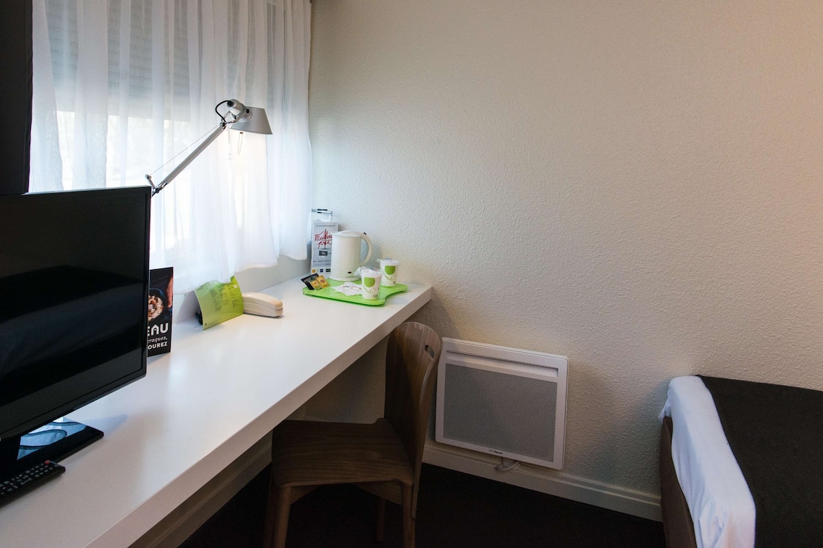 Une chambre d’hôtel équipée d’un bureau, d’un ordinateur, d’une lampe, d’une chaise, d’un lit et d’une fenêtre avec des rideaux.