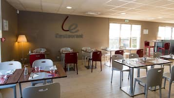 Ravintolassa on pöytiä ja tuoleja, seinässä on kyltti, jossa lukee ”Le Restaurant”, ja ikkunasta tulee sisään luonnonvaloa.