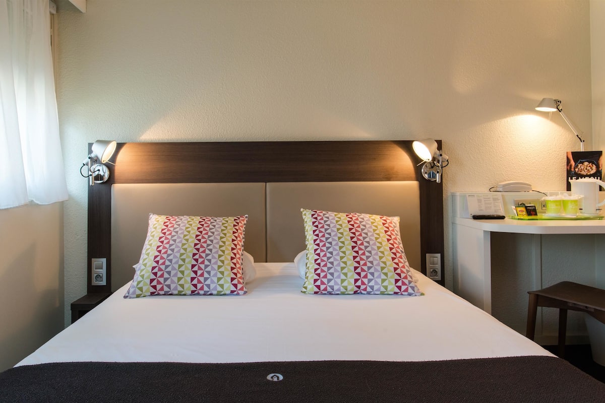 Une chambre d’hôtel avec un grand lit, deux coussins à motifs, une tête de lit agrémentée de lampes murales, une table de chevet avec une lampe, un petit bureau avec un téléphone et un menu, et une fenêtre avec des rideaux légers.