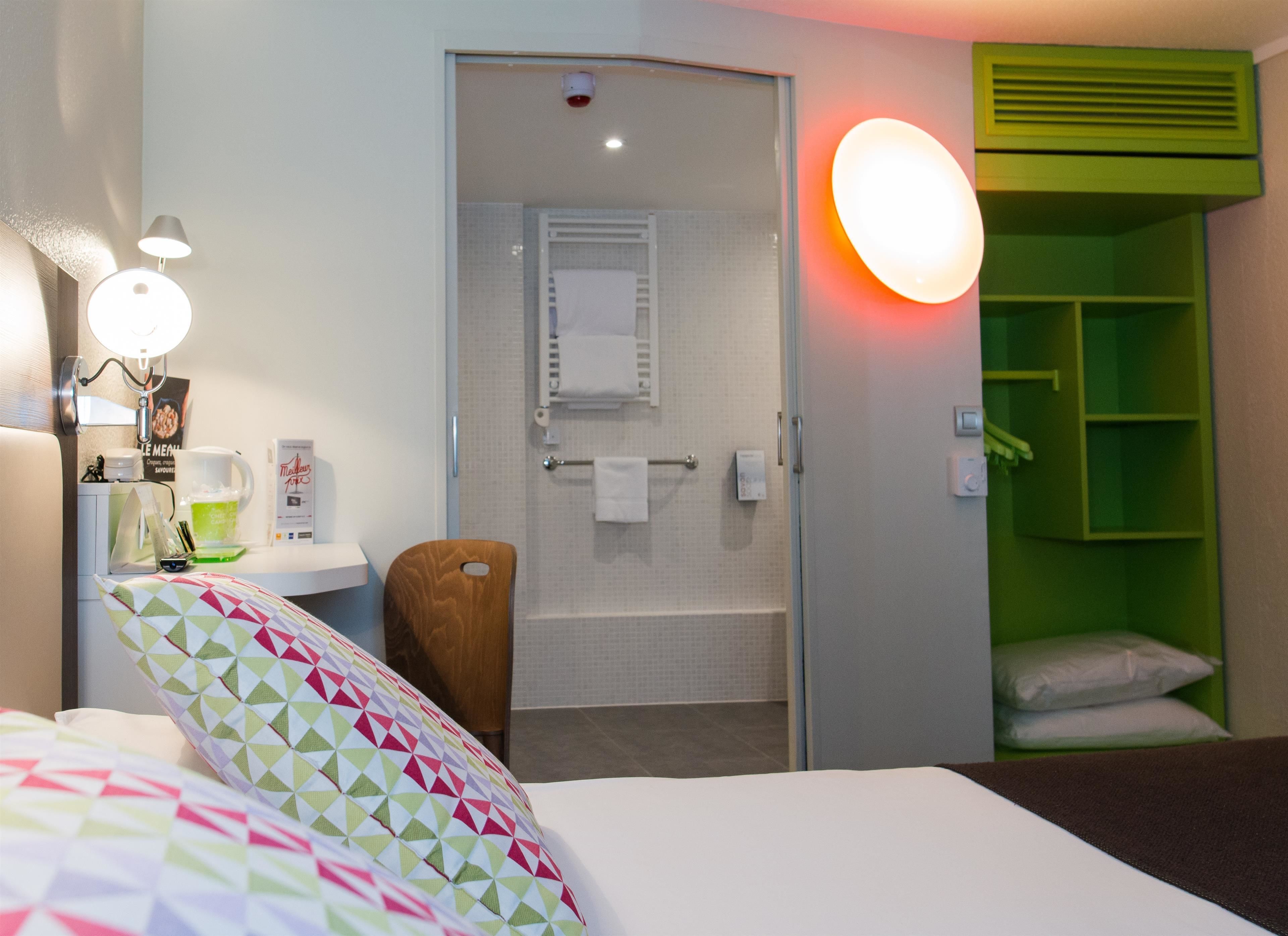 Ein Hotelzimmer mit einem Bett, einem Sessel, einem kleinen Tisch mit Lampe, einem grünen Wandschrank und einem Badezimmer mit Dusche und Handtuchhalter.