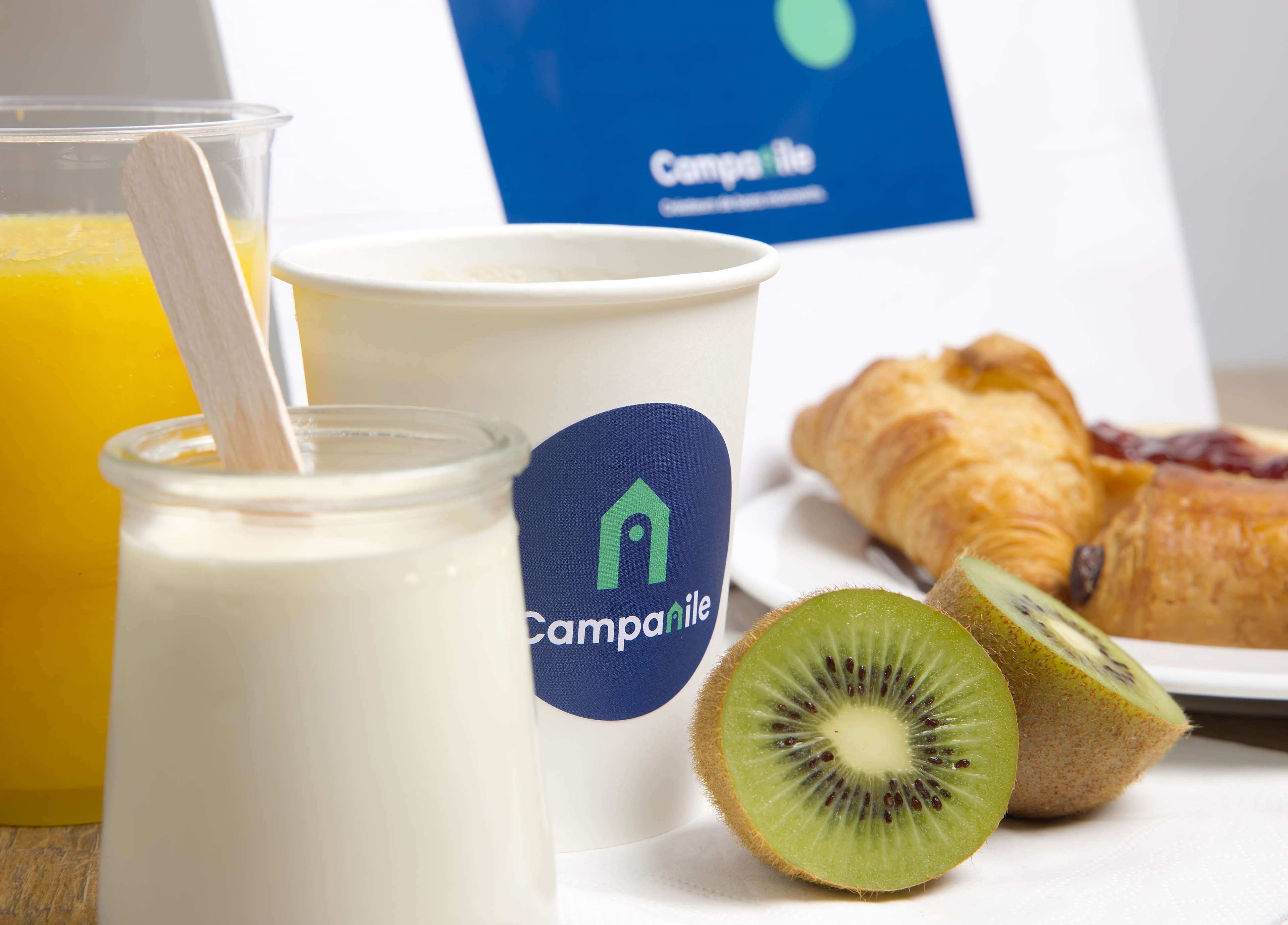 A breakfast setting with a glass of orange juice, a jar of yogurt, a cup with a logo, a croissant, and a kiwi slice.