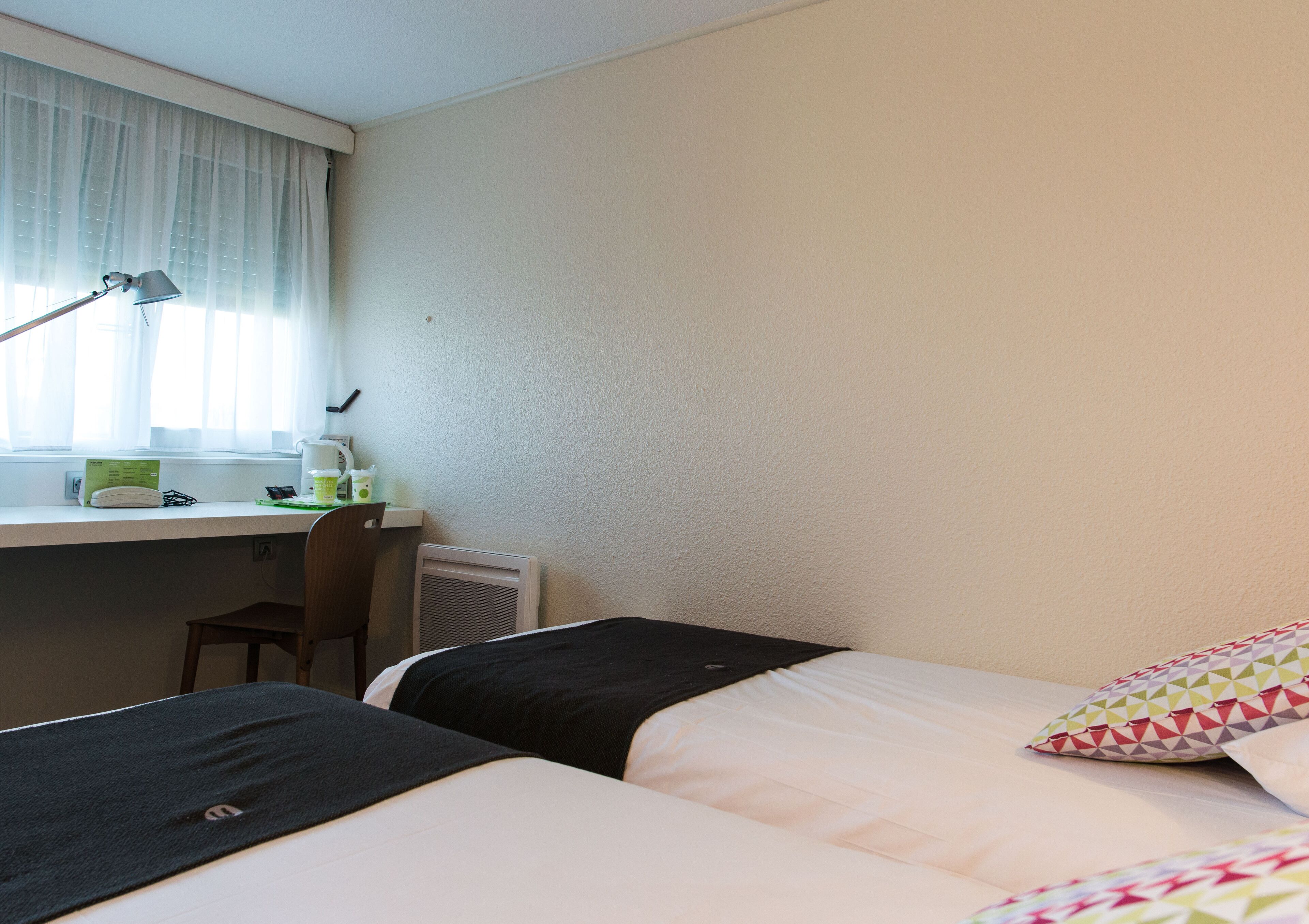 Habitación de hotel con dos camas, un escritorio con silla, una lámpara y una ventana con cortinas.