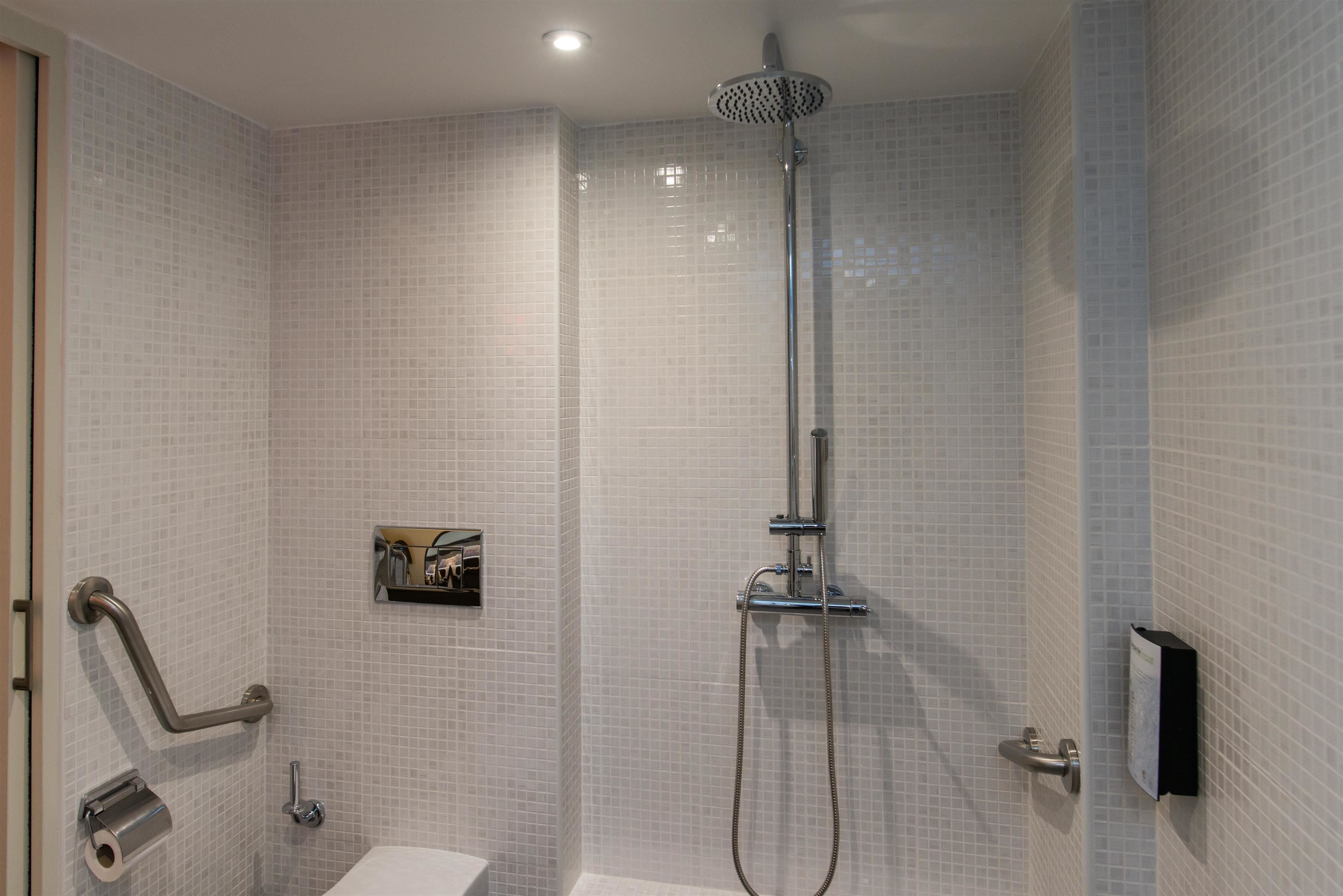 Ett badrum med vita kakelbeklädda väggar, en dusch med ett justerbart duschmunstycke, en toalett och ett handtag för att underlätta användningen för personer med funktionsnedsättning.