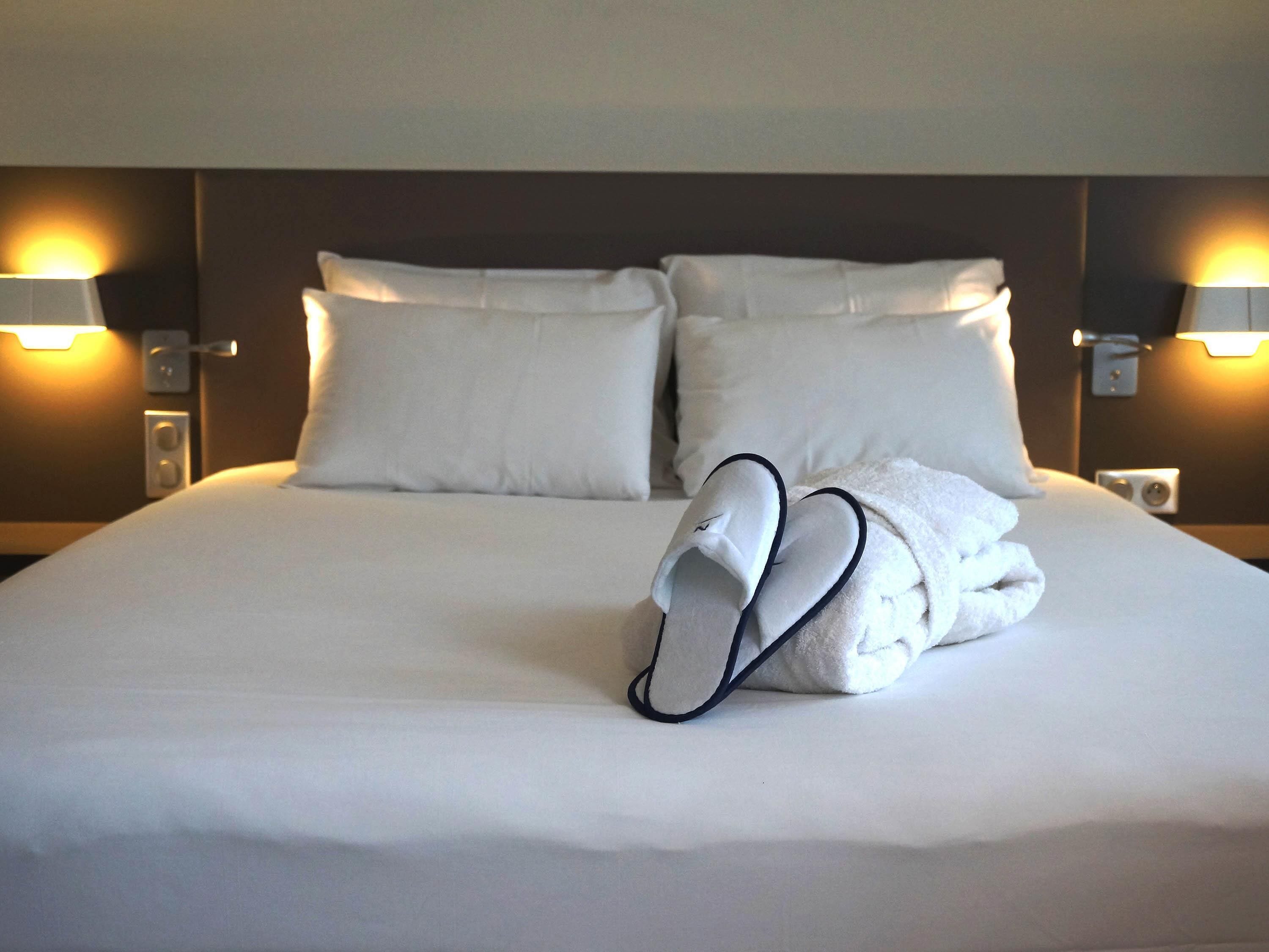 A neatly made bed with white linens and a folded towel on top. There are two wall-mounted lamps on either side of the bed.