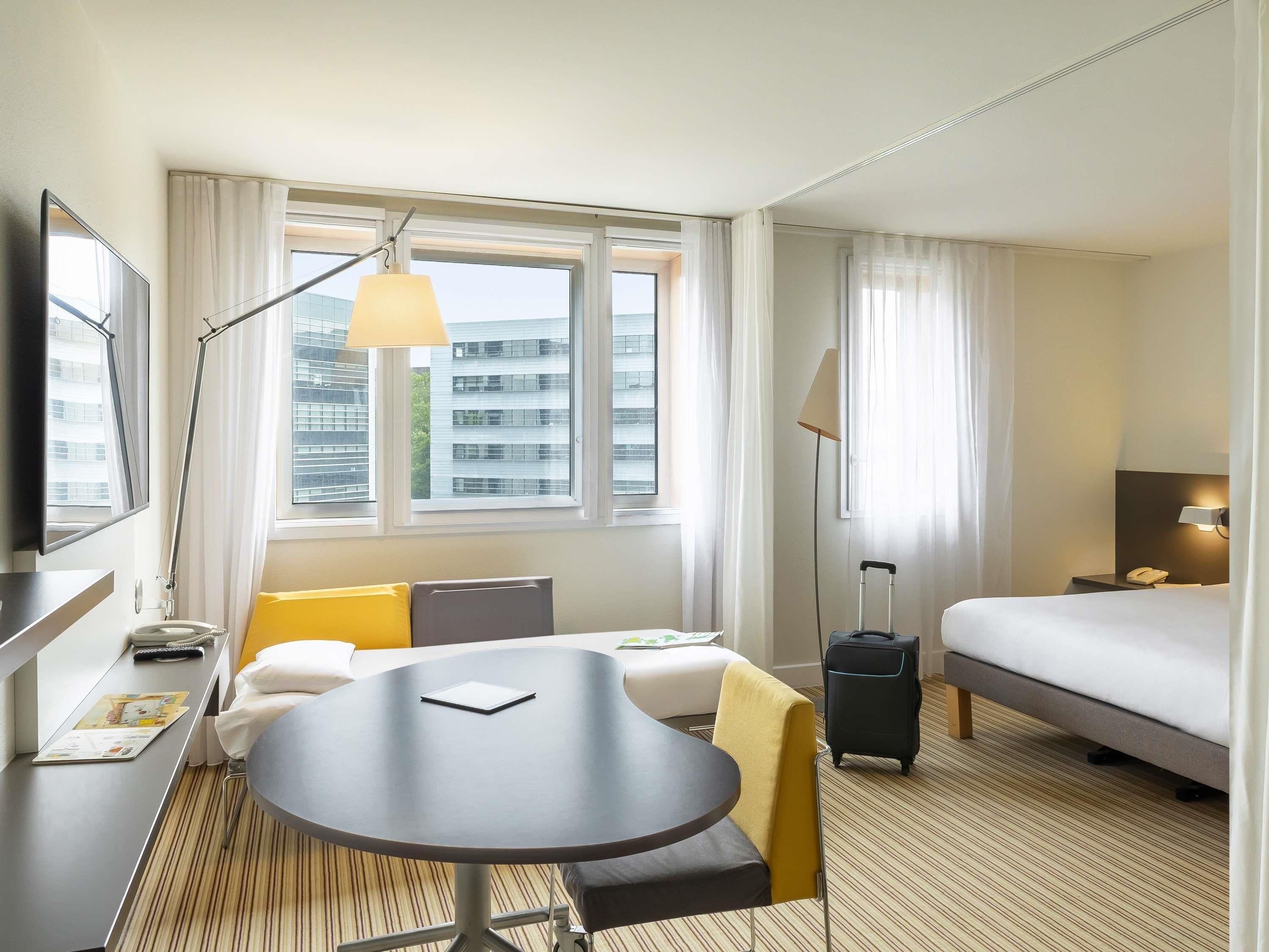 A hotel room with a bed, a desk with a lamp, a chair, a table, and a view of buildings through the window.