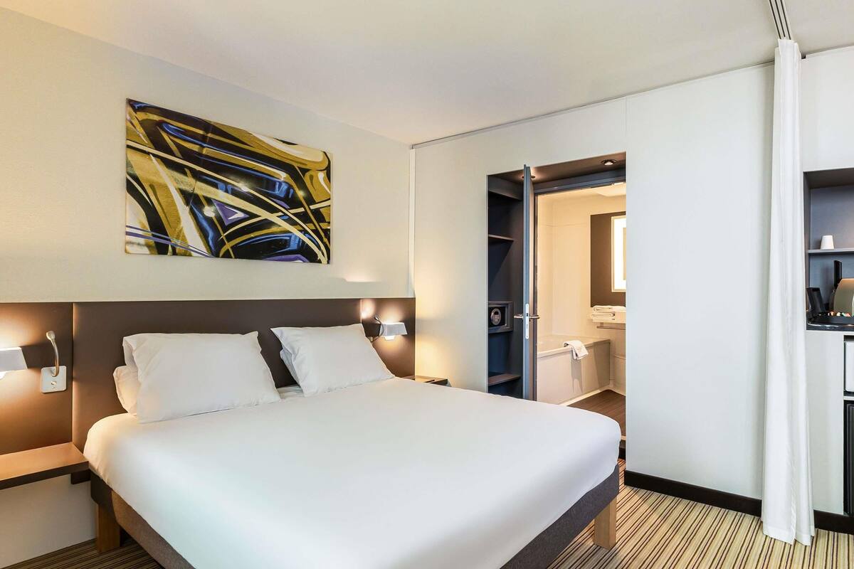 Une chambre d’hôtel dotée d’un grand lit, de tables de chevet, d’un tableau abstrait accroché au mur, d’un dressing et d’une salle de bain avec baignoire et porte-serviettes.