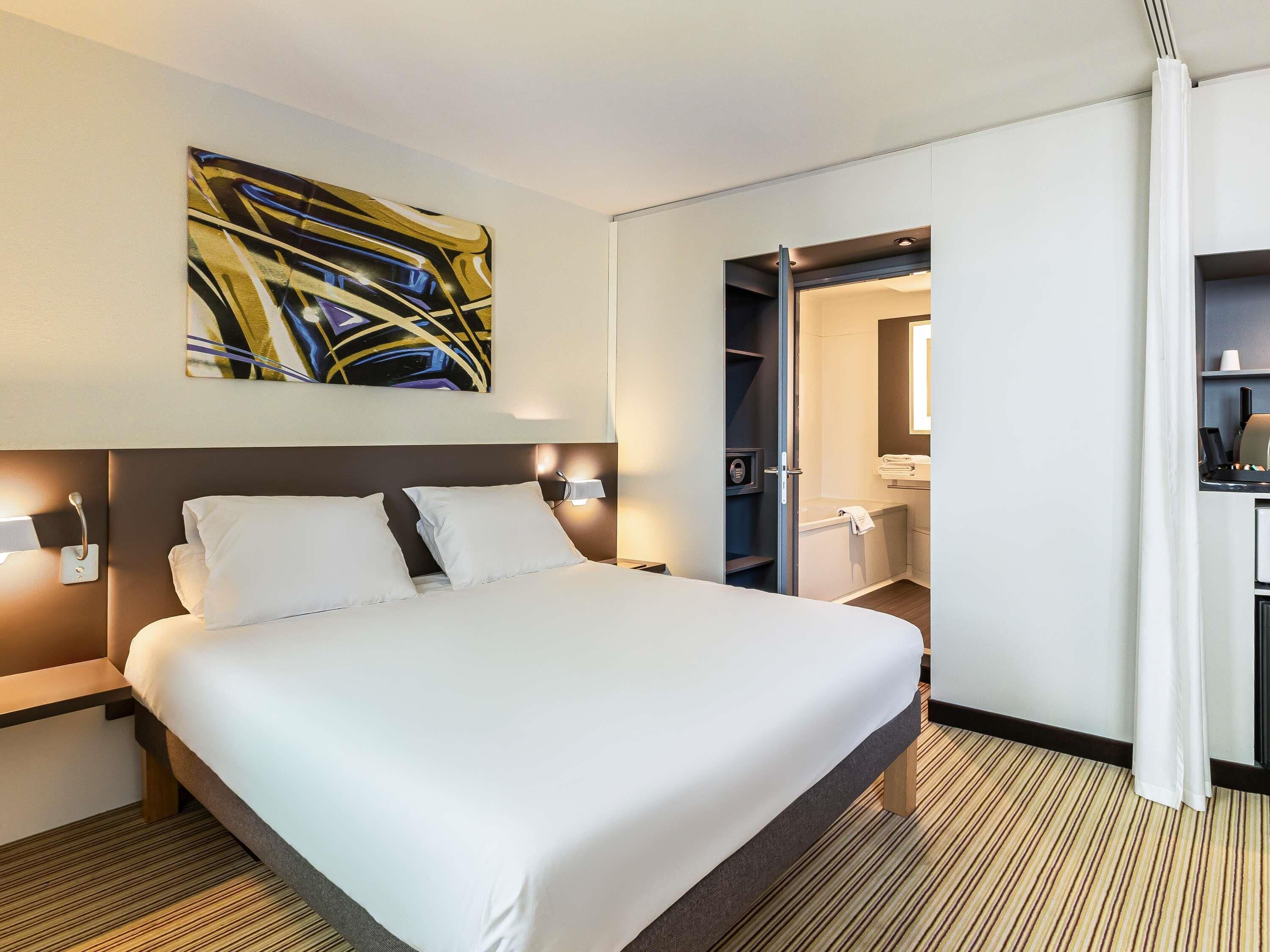 Habitación de hotel con una cama grande, un cuadro en la pared, un baño visible a través de una puerta abierta y un pequeño escritorio.
