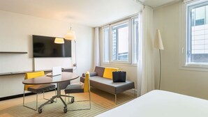 Habitación de hotel con cama, escritorio con silla, sofá con cojines, televisor montado en la pared y ventanas amplias.