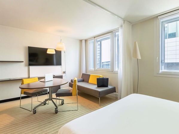 Habitación de hotel con cama, escritorio con silla, sofá con cojines, televisor montado en la pared y ventanas amplias.