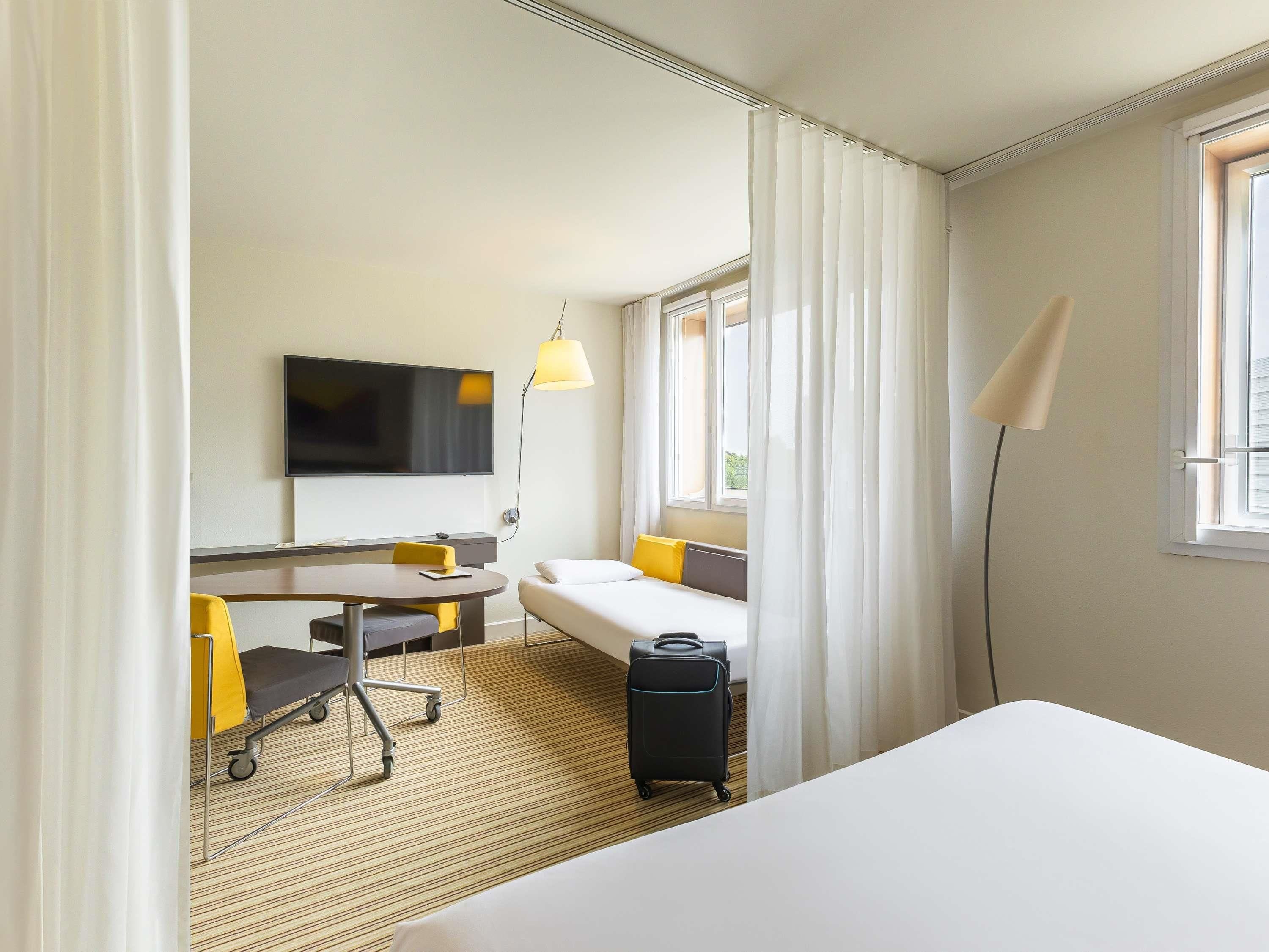 A hotel room with a bed, a desk with a chair, a TV, a lamp, a suitcase, and a window with curtains.