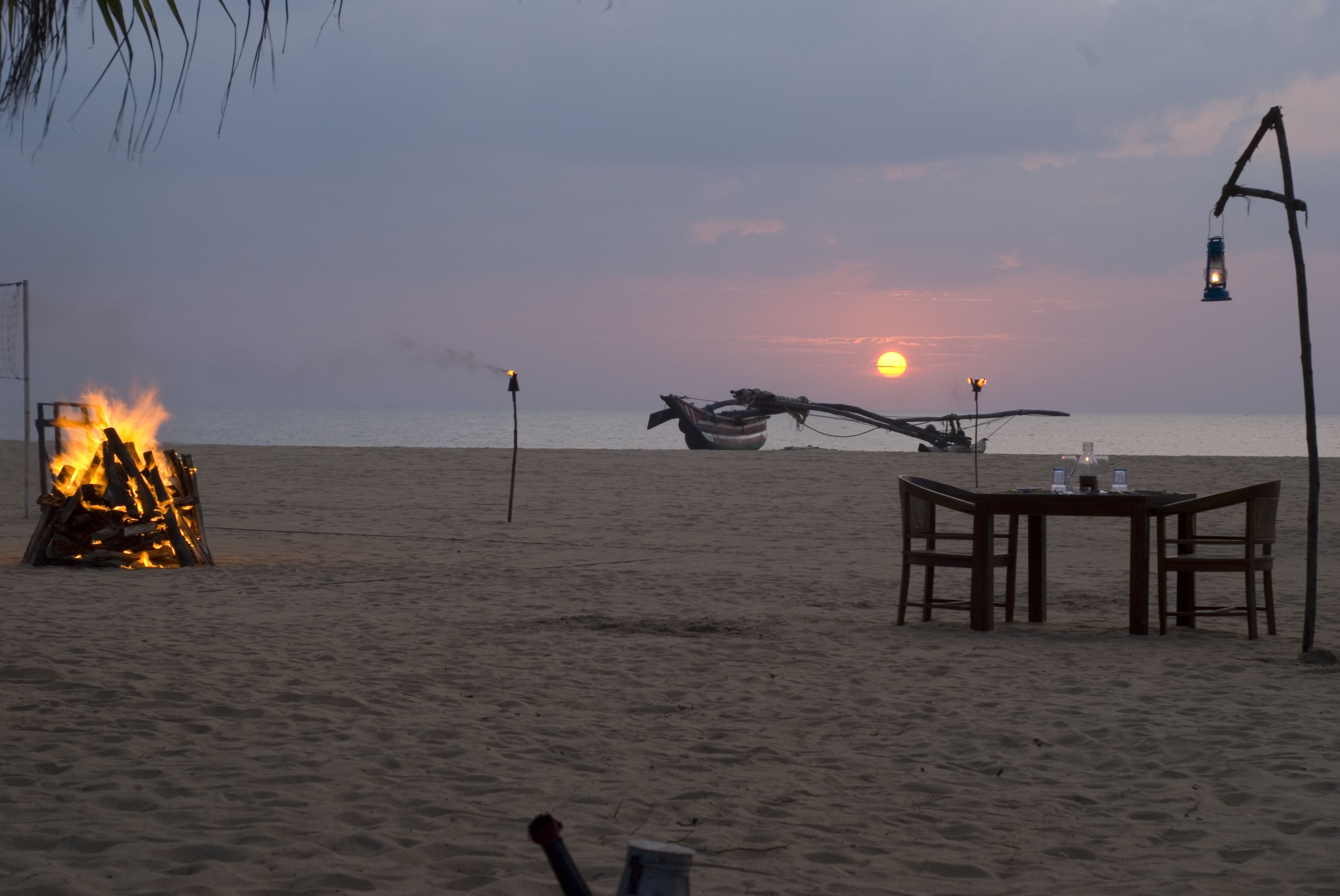 A beach scene with a fire, a table with chairs, and a sunset in the background.