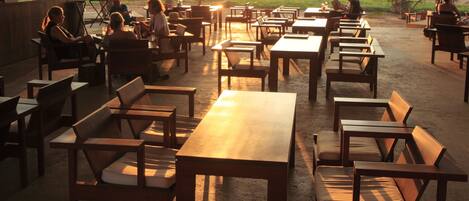 Un espace extérieur aménagé avec des tables et des chaises, des palmiers et une vue sur la plage au coucher du soleil.