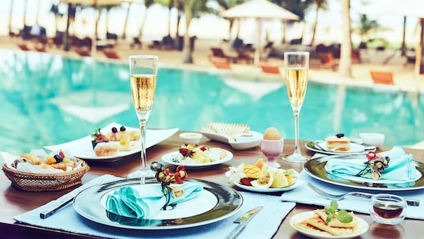 A dining table with plates of food, glasses of champagne, and a poolside setting with umbrellas and lounge chairs. - Jetwing Beach (Negombo)