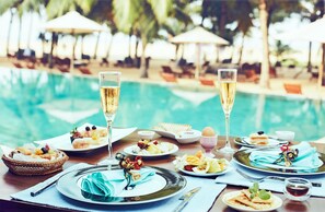 A dining table with plates of food, glasses of champagne, and a poolside setting with umbrellas and lounge chairs. - Jetwing Beach (Negombo)
