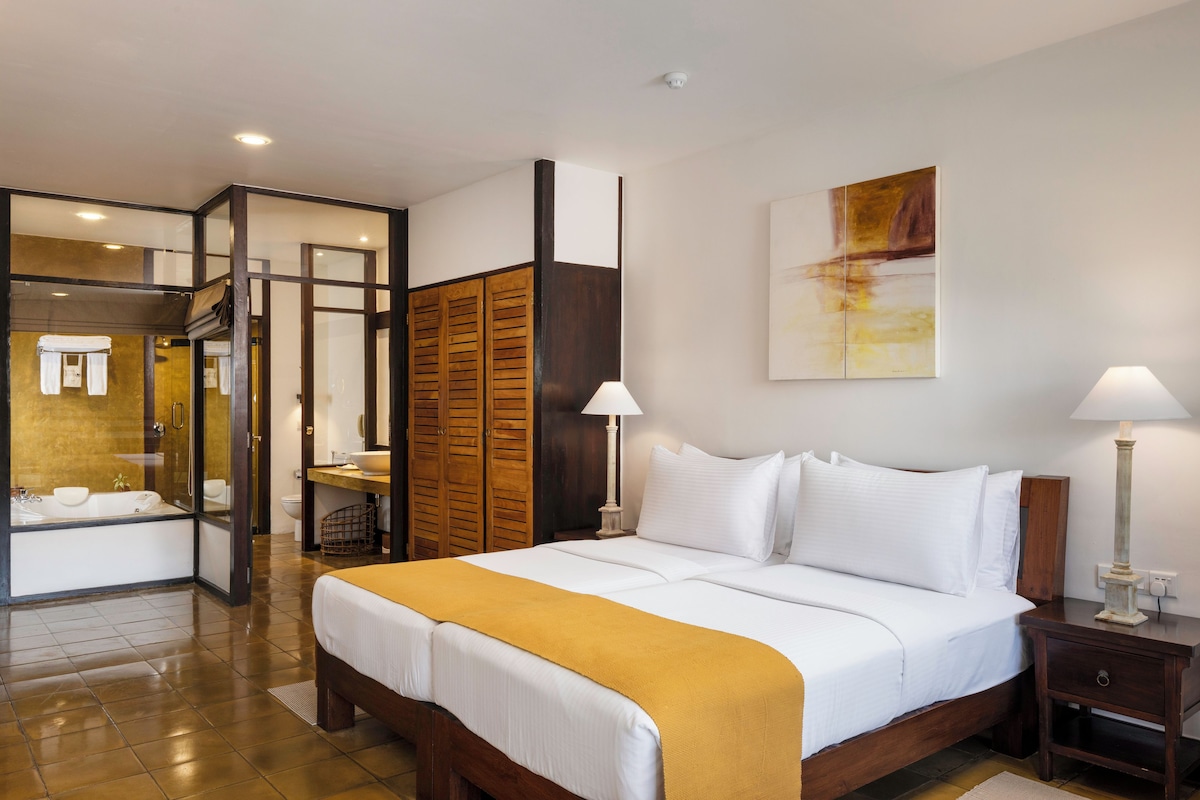 A hotel room with a large bed, bedside lamps, a wooden wardrobe, a bathroom with a bathtub, and a large window.