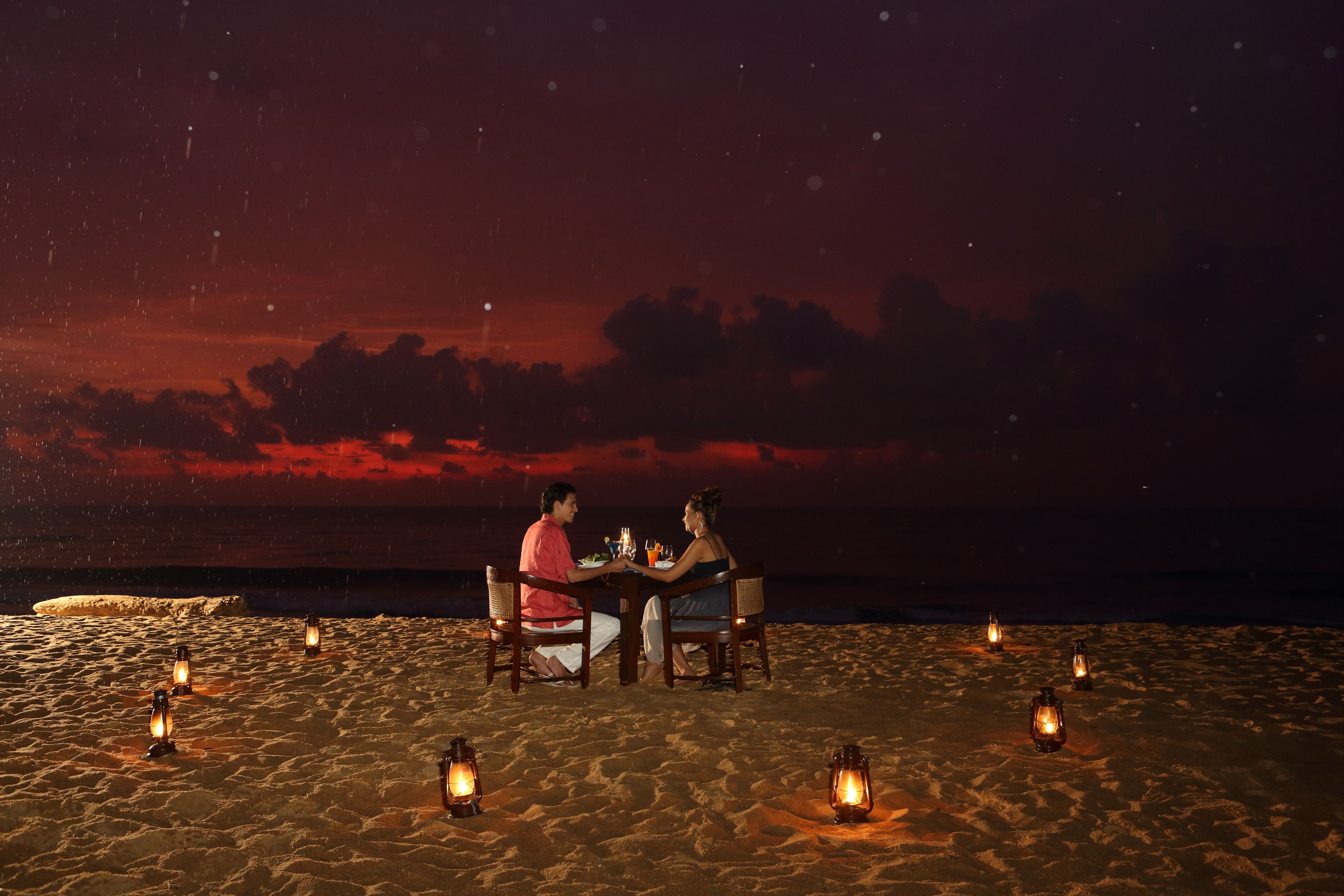 Una pareja cenando en una mesa en la playa de noche, con faroles alrededor.