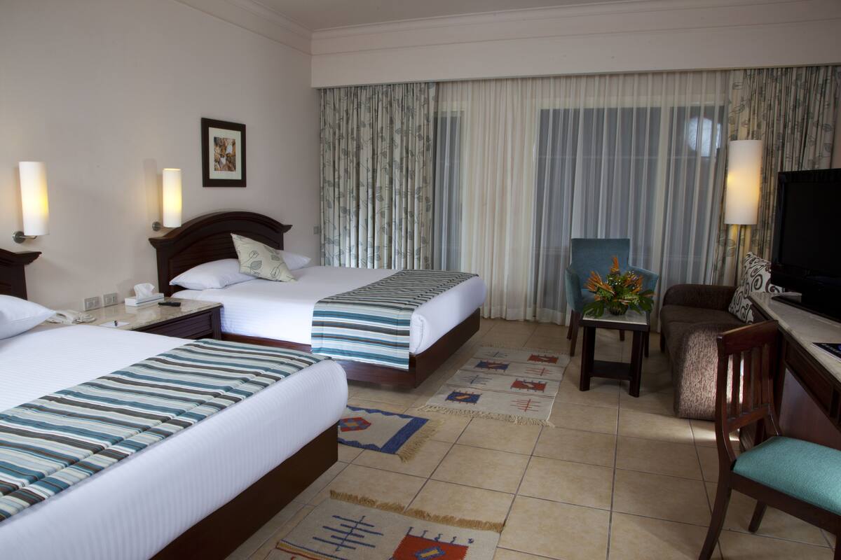 A hotel room with two beds, a desk, a chair, a TV, and a large window with curtains.