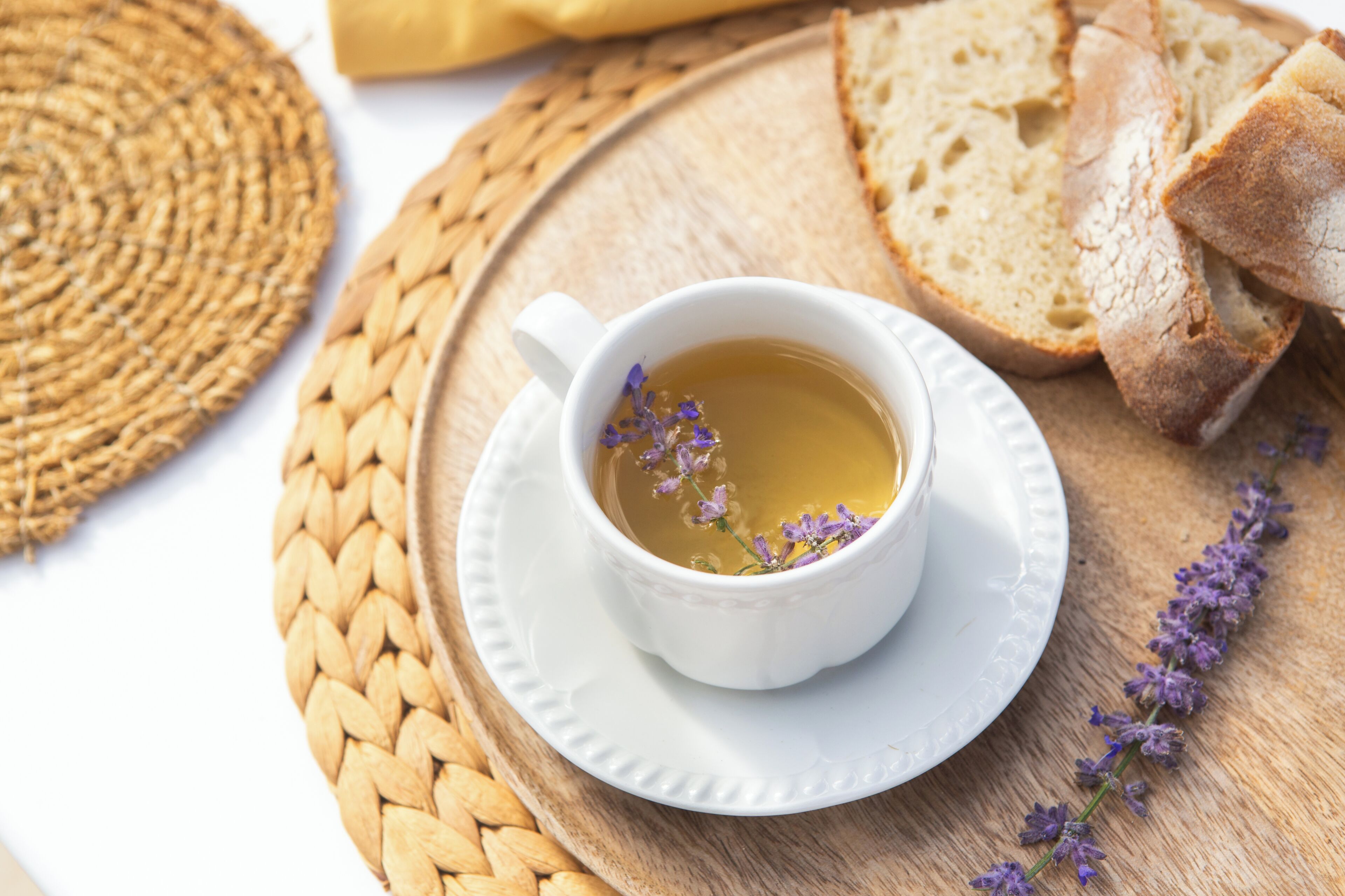 A cup of tea with lavender on a woven mat, accompanied by slices of bread.