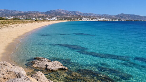 Una playa costera con aguas turquesas cristalinas, costa rocosa y montañas con edificios blancos al fondo.