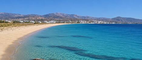 A coastal beach with clear turquoise water, rocky shoreline, and a backdrop of mountains and white buildings.