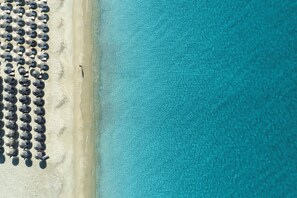 Eine Vogelperspektive zeigt einen Strand mit Reihen von Sonnenschirmen und einem klaren, blauen Meer.