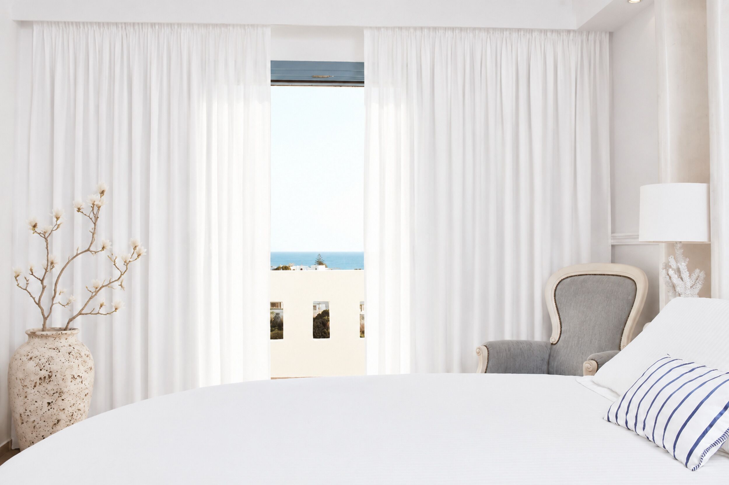Une chambre d’hôtel avec un grand lit, une chaise, un vase de fleurs séchées et une vue sur la mer à travers de fines rideaux.