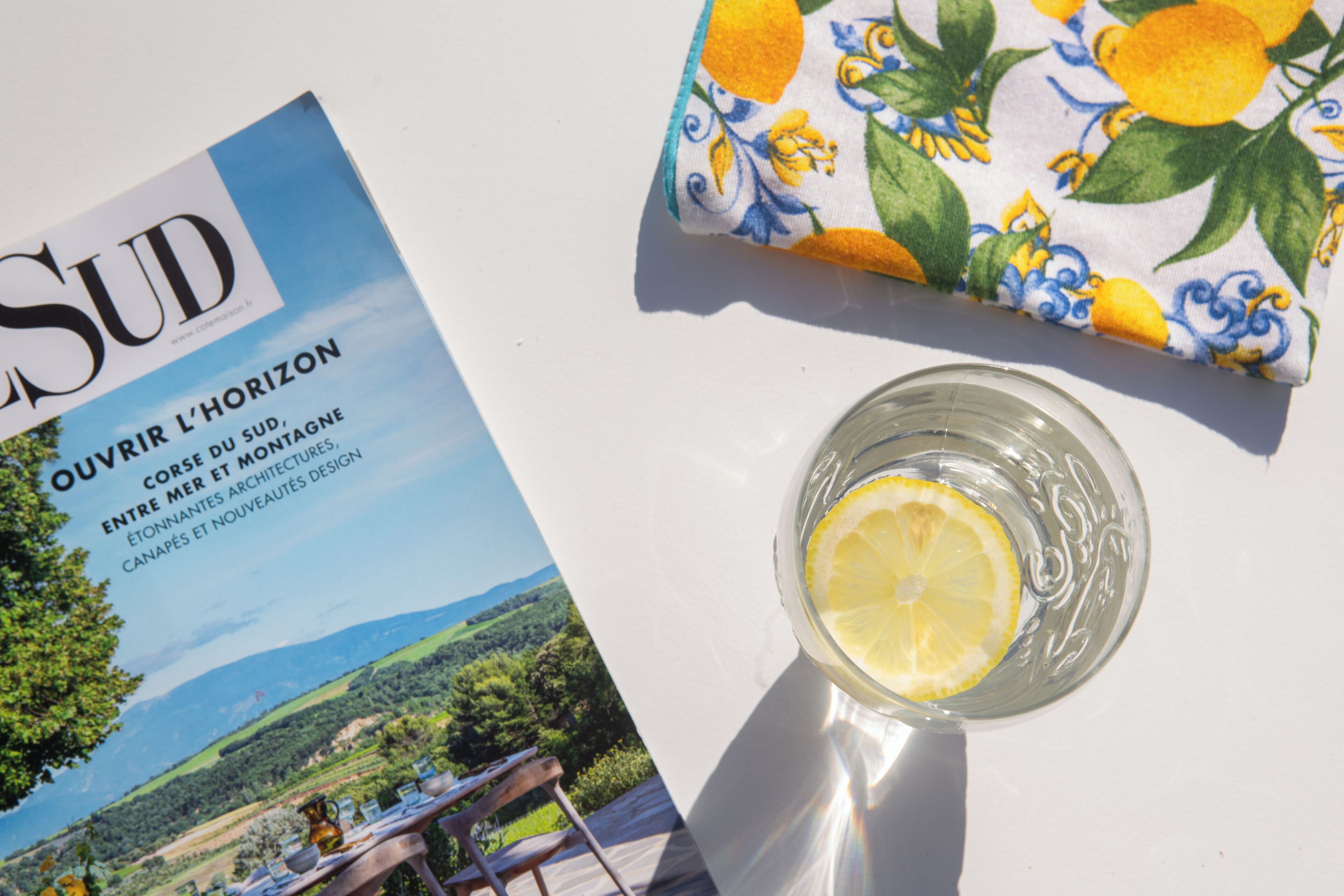 En tidning med titeln ”SUD” som visar ett landskapsfoto, ett glas med en citronskiva och ett bord med en duk på.