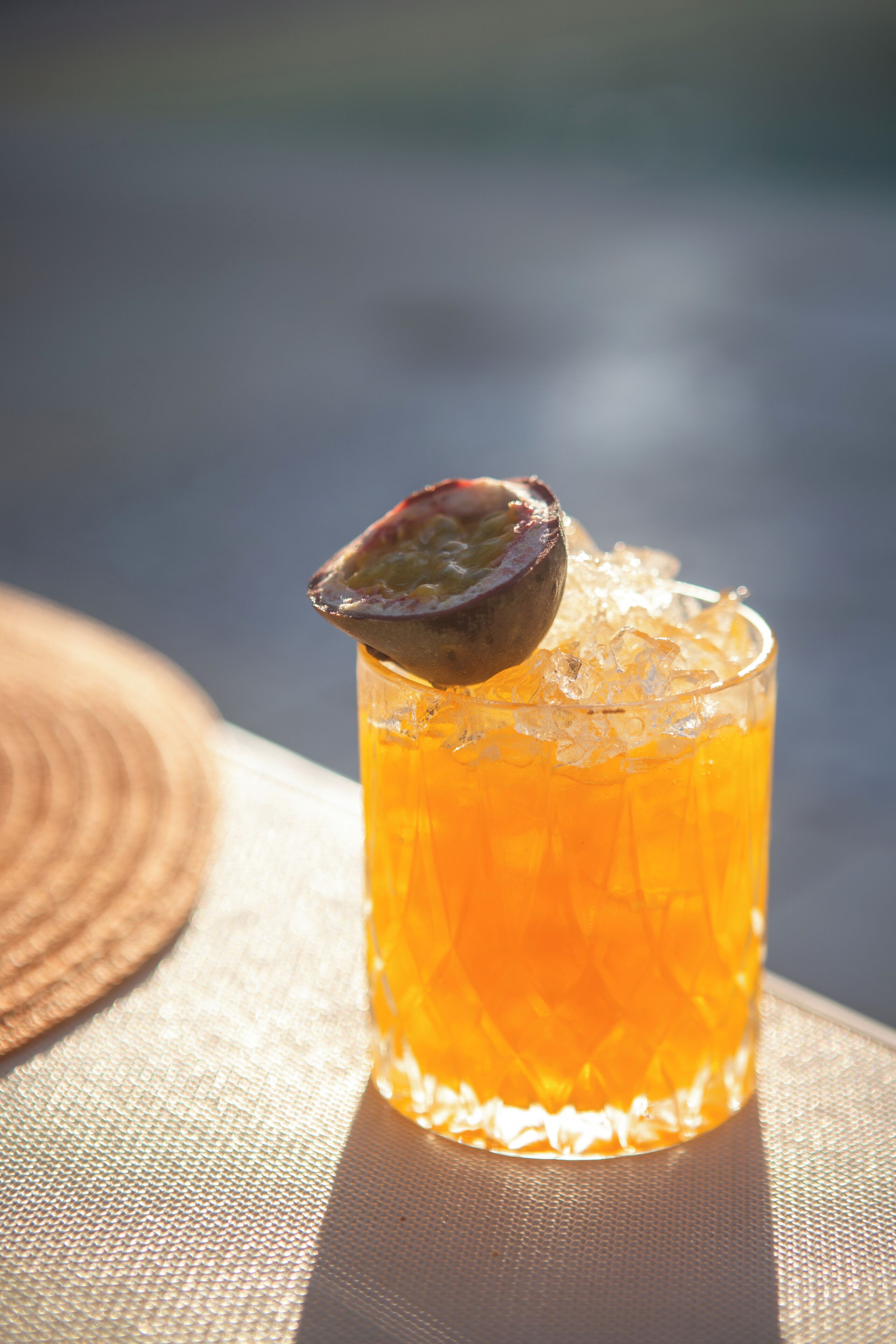A glass of amber-colored beverage with ice and a slice of fruit on a table.