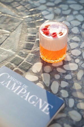 A glass of beverage on a textured surface with a book titled 'Maison de Campagne' beside it. - Lagos Mare Hotel (Naxos)