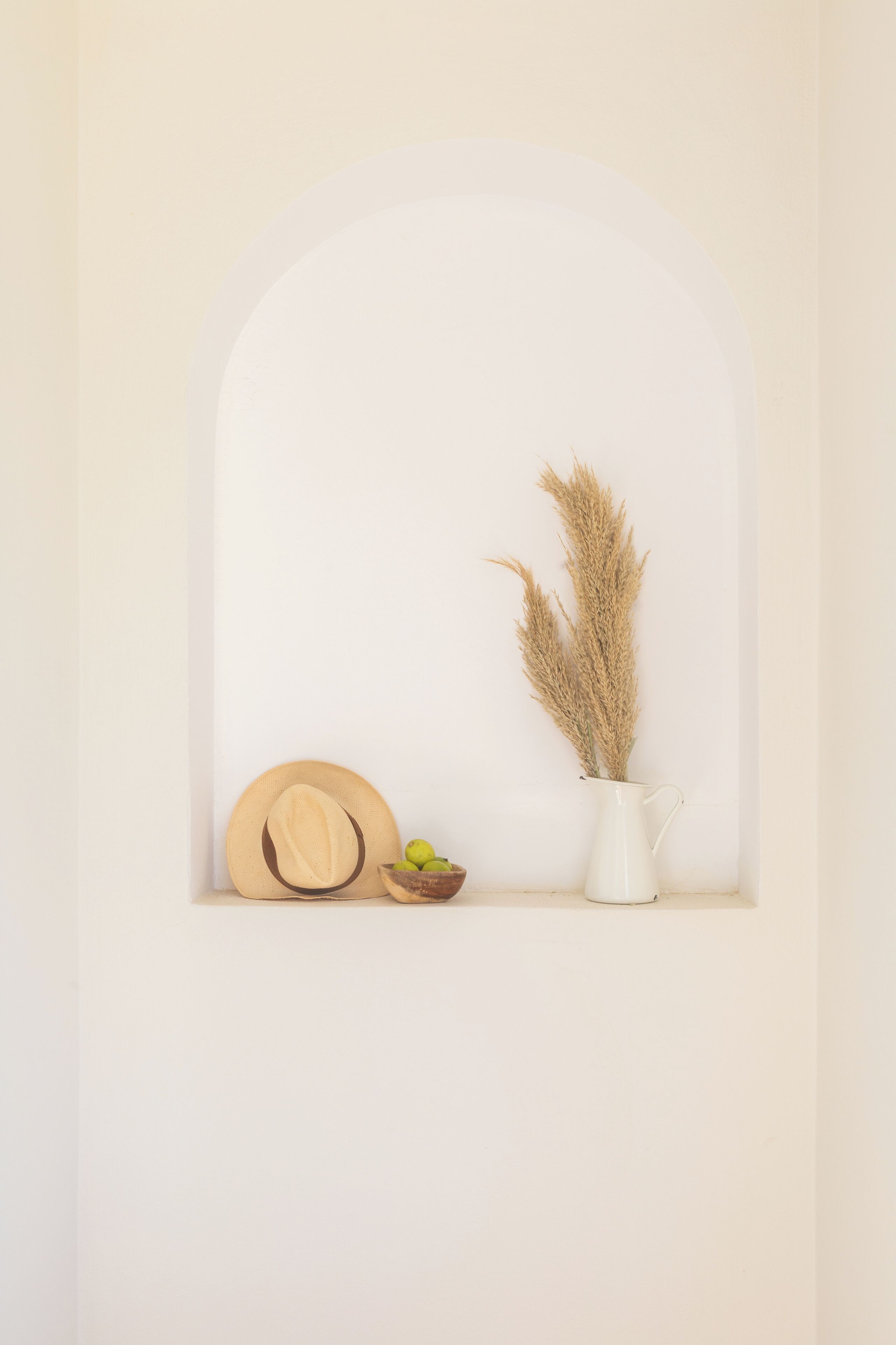 A white arched alcove with a wooden hat, a small bowl, and a white vase with pampas grass.
