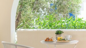 Una mesa con alimentos para el desayuno y una planta en maceta, sobre un fondo de follaje y una pared blanca.