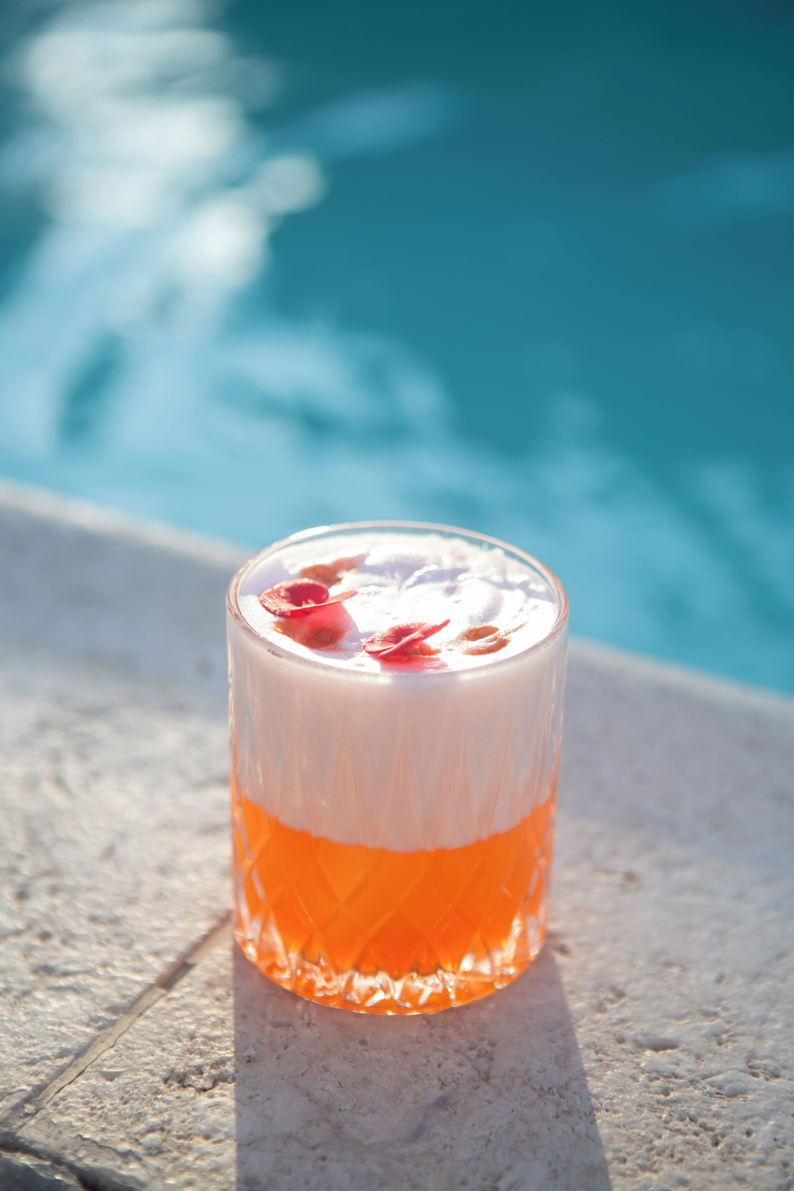 A glass with a layered drink, placed on a stone surface near a pool.