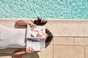 A person lying on a stone ledge by a pool, holding a magazine. - Lagos Mare Hotel (Naxos)