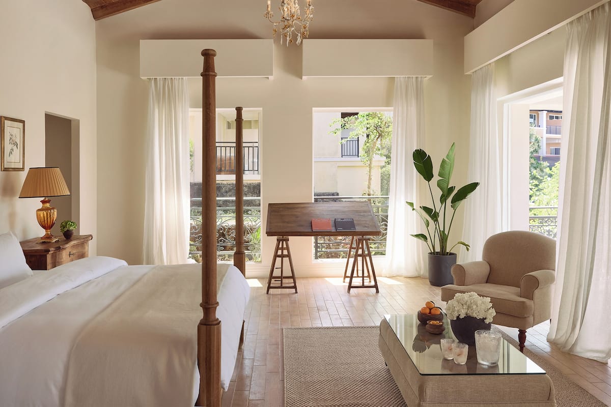 Um quarto espaçoso com uma cama grande, teto de madeira, lustre, varanda com vista e um espaço de estar com mesa e cadeira.