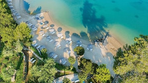 Vista aérea de una playa con filas de tumbonas y sombrillas, agua turquesa cristalina y barcos a lo lejos.